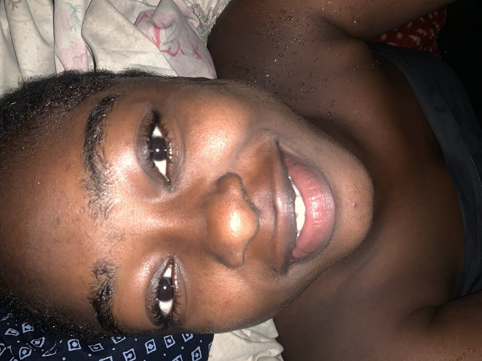 The image is a close-up, low-angle portrait of a young Black individual, likely female, who appears to be lying down. The subject's face is the primary focus, with her eyes open and looking towards the camera. She has dark skin, thick eyebrows, and dark eyelashes. Her lips are slightly parted, revealing white teeth. There are visible water droplets on her forehead, cheeks, and neck, suggesting she may be sweating or has recently been in contact with water.
The background is a mix of textures and patterns. To the left, a dark blue fabric with a white geometric pattern is visible. To the right and behind the subject's head, a lighter-colored fabric with a floral pattern is draped. The lower part of the image shows a dark garment, possibly a top or dress, and a glimpse of a red patterned fabric beneath.
The lighting is dim, creating a sense of intimacy and potentially indicating that the photo was taken indoors or at night. The overall impression is of a casual, personal moment captured in what appears to be a domestic setting.
The prompt asks for the location in the city of Rafin Par, Nigeria. However, the image itself provides no visual cues or information that would allow for the identification of a specific location within Rafin Par, Nigeria. The scene depicted is generic and could be found in many places. Therefore, it is not possible to provide details about the location in Rafin Par based solely on the image.