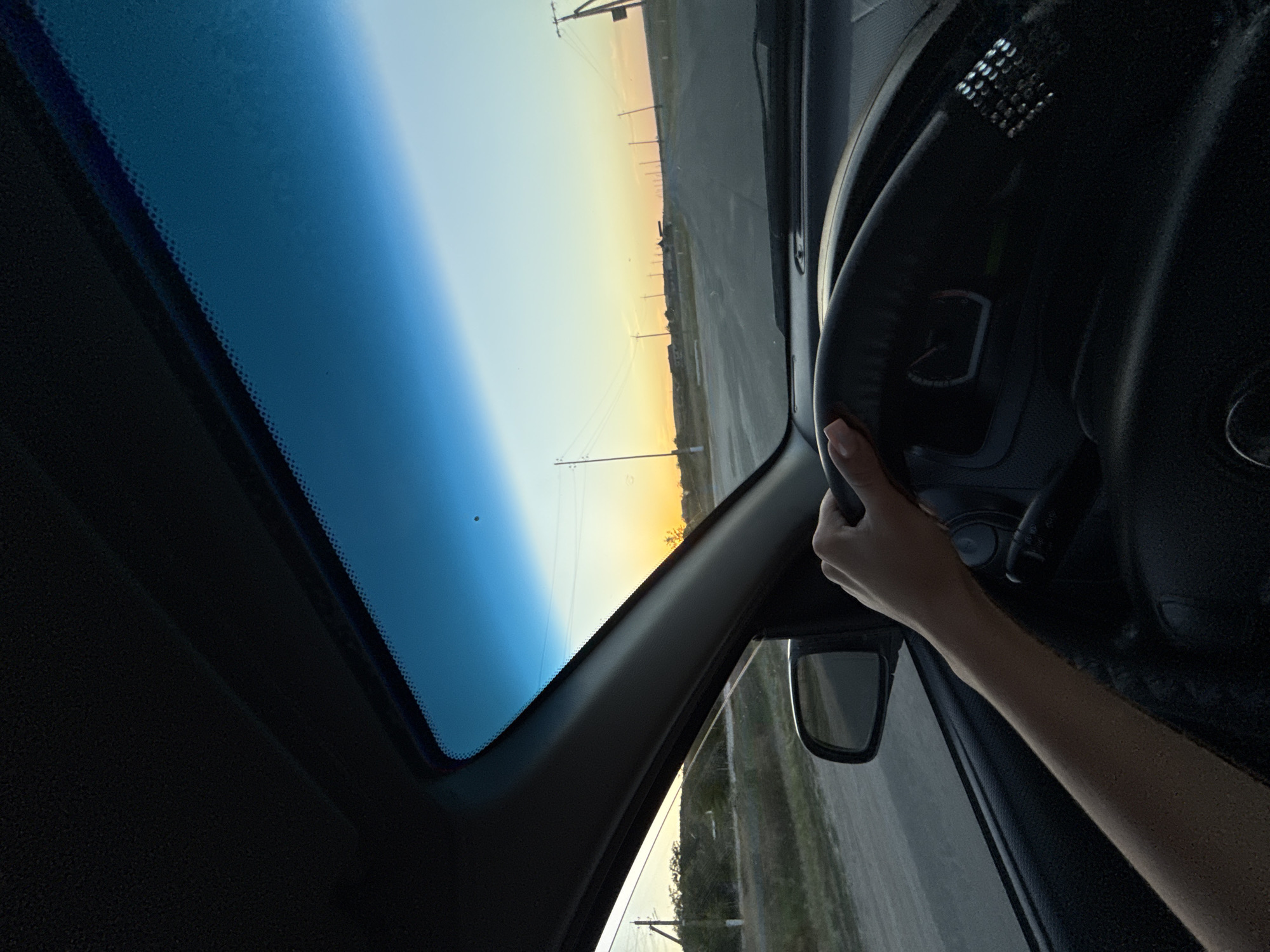 An over-the-shoulder shot from inside a vehicle shows a person's left arm and hand gripping the steering wheel. The person is driving on a paved road in a rural area, likely in or near Cioc-Maidan, Moldova, during sunset. Power lines and sparse vegetation line the road, which stretches into the distance towards a horizon illuminated by the setting sun. The sky transitions from a deep blue at the top to a soft orange and yellow hue near the horizon. The interior of the car, including the dashboard and windshield, is visible.