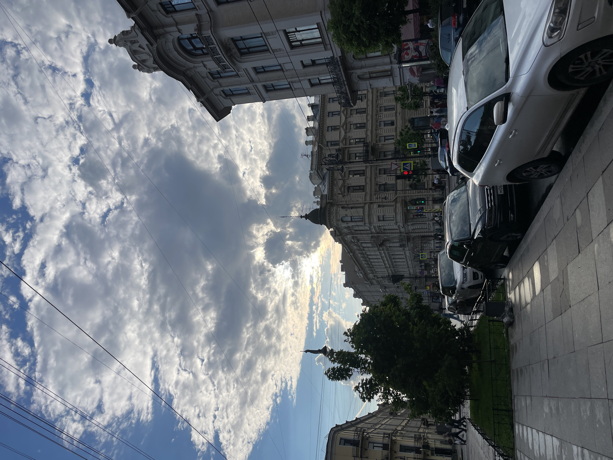 The image depicts a vibrant street scene in what appears to be a historic European city, confirmed by context as Saint Petersburg, Russia. The perspective is from ground level, looking down a street lined with ornate, multi-story buildings of classical architectural styles.
The sky dominates the upper portion of the frame, showcasing a dynamic interplay of bright blue patches and dramatic, voluminous white and grey clouds. The sun, partially obscured by clouds, casts bright highlights and long shadows, suggesting late afternoon or early evening. Numerous overhead wires crisscross the sky, indicative of urban infrastructure, possibly for trams or trolleybuses.
On the right side of the street, cars are parked along the curb. A prominent white SUV is visible in the foreground, with part of its license plate, "M06," clearly legible. Behind it are several darker vehicles. The paved sidewalk next to the parked cars appears damp, possibly from recent rain, reflecting the light.
Further down the street on the right, traffic signals, including a traffic light and a yellow triangular pedestrian crossing sign, are visible. Buildings on this side of the street also feature intricate detailing. Scattered pedestrians can be seen in the distance along the sidewalk.
The left side of the image features more impressive architecture, including a building with a decorative façade and a distinctive dome or spired feature silhouetted against the bright sky. Green trees with lush foliage are present, both along the sidewalk and amidst the buildings, adding a touch of nature to the urban environment. A small patch of green grass is visible along the left sidewalk in the foreground.
The overall impression is one of an active city street under a dramatic sky, blending historical grandeur with contemporary urban life.