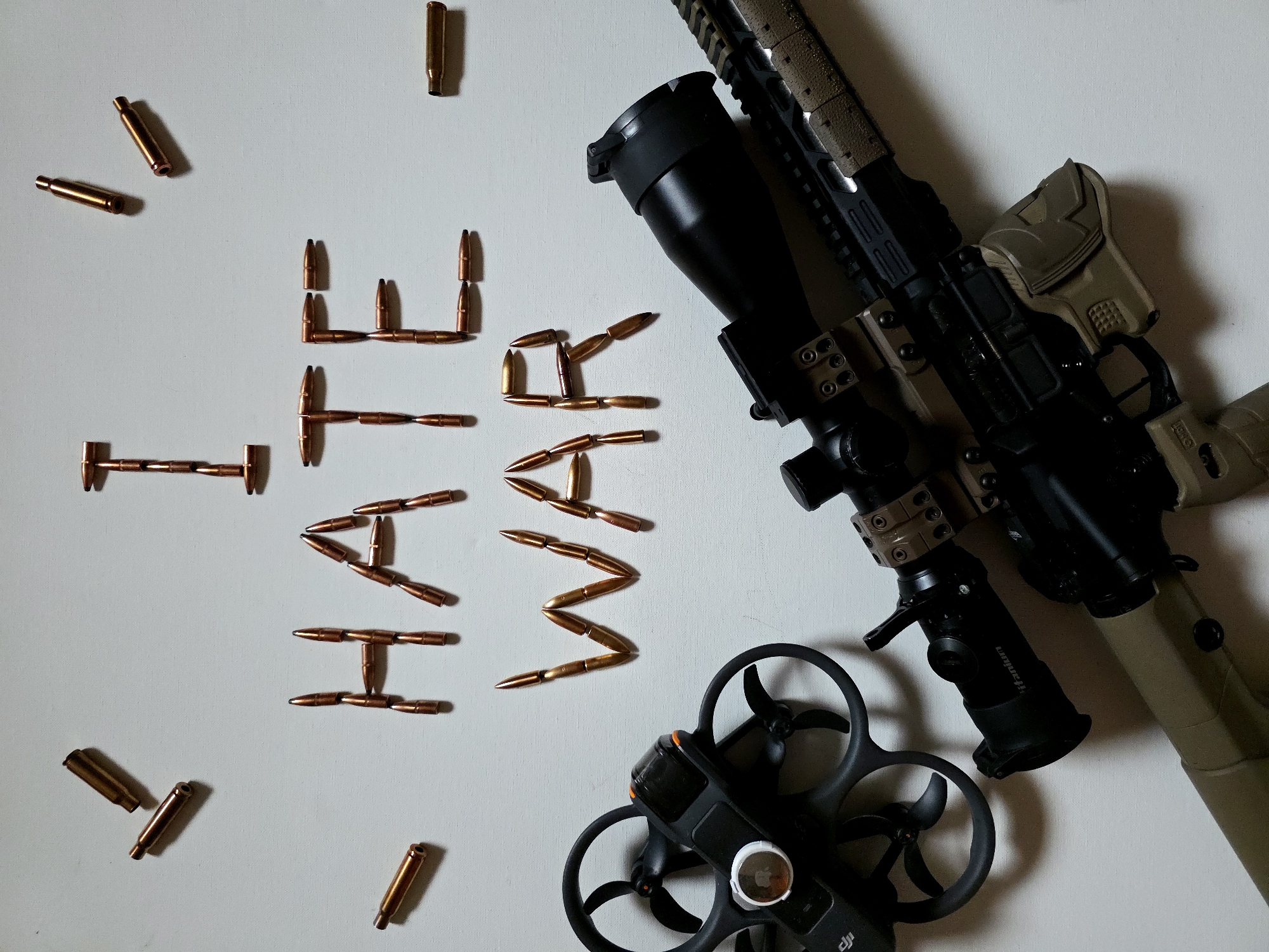 An overhead shot presents a collection of objects arranged on a stark white surface, conveying an anti-war sentiment. A modern rifle, predominantly tan and black, with a large black scope, is positioned diagonally across the upper right side of the frame. Below the rifle, towards the bottom right, sits a black DJI drone with its four propellers visible.
Numerous bullet projectiles are artfully arranged in the central and left portions of the image to spell out the words "HATE WAR" in capital letters. "HATE" is on the left, formed by individual projectiles, and "WAR" is on the right, similarly constructed. Several loose bullet casings and projectiles are scattered around the periphery of the white background.
The scene appears to be an indoor setup, likely staged, with a clear white backdrop and soft, indirect lighting that creates subtle shadows. The juxtaposition of the rifle, bullets, and drone—representing instruments of conflict and modern warfare technology—with the explicit "HATE WAR" message creates a powerful visual commentary. The brand "DJI" is visible on the drone, and the word "tantalum" can be seen on an accessory attached to the rifle's scope. No individuals are present in the image.