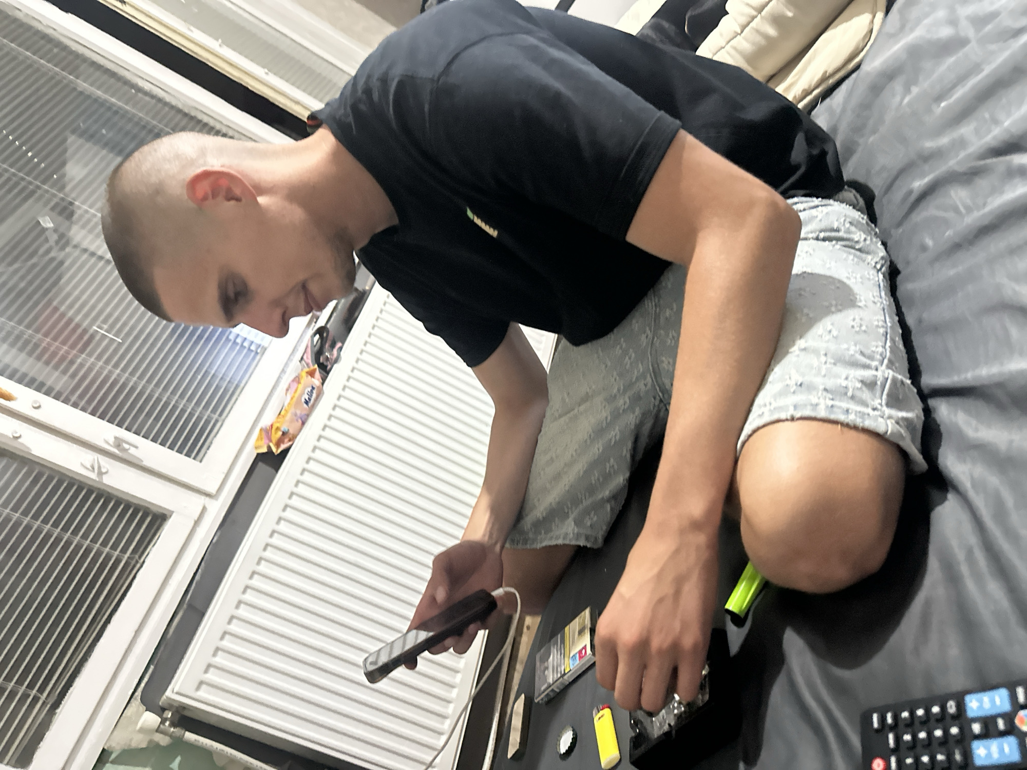 The scene, located in Sofia, Bulgaria, depicts an adult male seated on a dark grey textile surface. He exhibits a closely cropped hairstyle and wears a dark-colored short-sleeved t-shirt and light-colored patterned shorts. He holds a dark smartphone connected by a white cable in his left hand, while his right hand is positioned near a black ashtray. Adjacent to the ashtray are a yellow disposable lighter, a pack of cigarettes displaying health warnings, and a silver bottle cap. A multi-buttoned black remote control is visible in the bottom left foreground. The background features a white-framed window equipped with horizontal blinds, situated above a white panel radiator. A section of light green wall is visible to the left of the radiator. A yellow and orange snack bag is present on a surface behind the radiator. The environment indicates an indoor residential setting.
