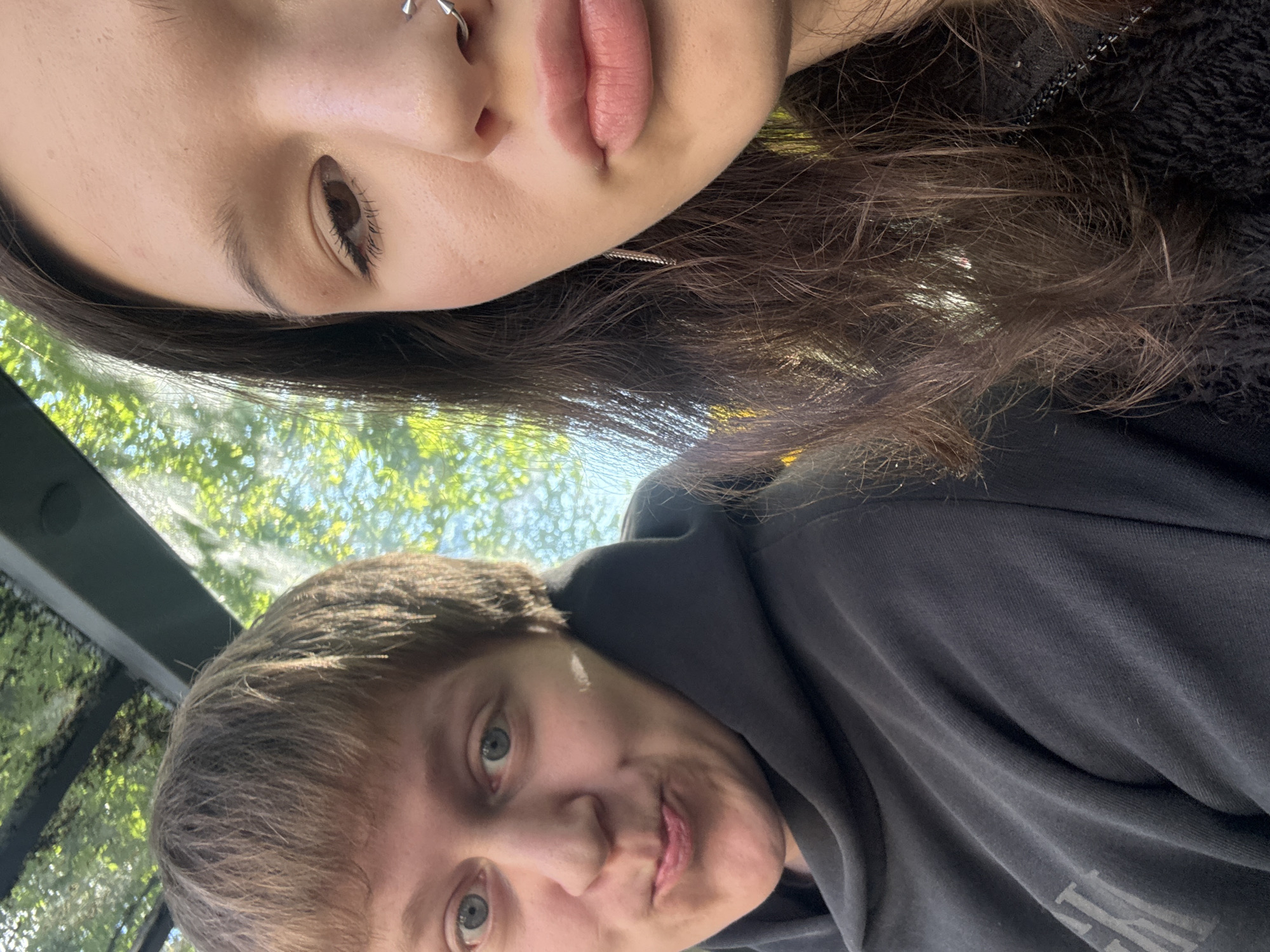 Two individuals are visible in the image. The individual at the top of the frame has dark brown hair, light skin, a nose ring, and is wearing what appears to be a dark-colored top. The person at the bottom of the frame has short, light brown hair, fair skin, and is wearing a gray hooded sweatshirt. The background includes foliage and a glimpse of what may be a glass structure or window reflecting the trees. The location within Severnaya Samarka, Russia, cannot be determined from the visual data provided.