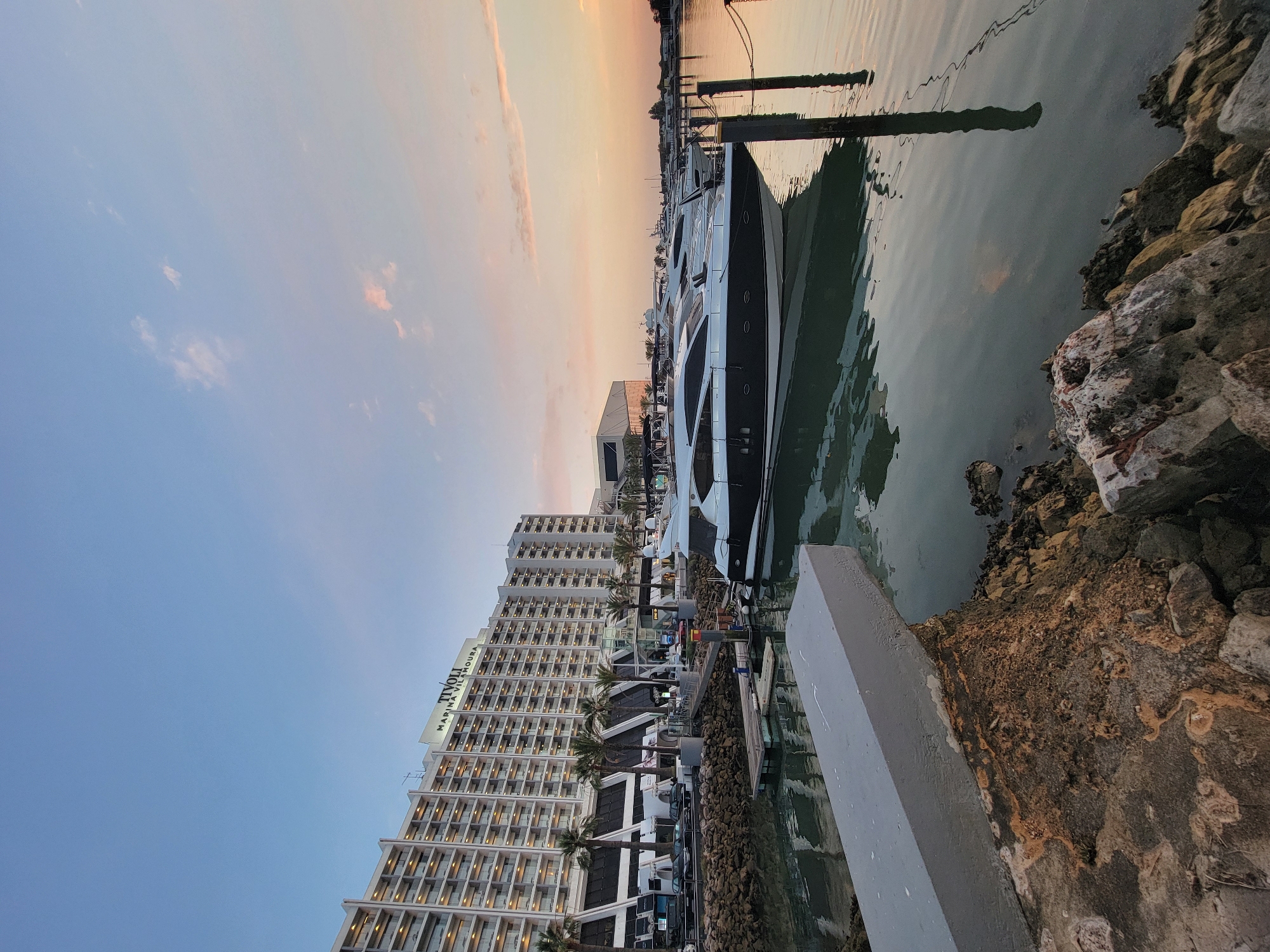 This is a photograph of the marina at Vilamoura, located in Quarteira, Portugal. The scene captures a waterfront view with a large hotel in the background and several yachts docked in the foreground. The hotel, identified by the "TIVOLI MARINA VILAMOURA" signage, is a multi-story building with a repetitive facade of balconies and windows, illuminated from within. Palm trees are visible along the waterfront in front of the hotel.
The marina itself is calm, with the water reflecting the soft colors of the sky and the boats. Two prominent yachts are the main subjects in the water. The closer yacht is a sleek, modern vessel with a dark hull and a white superstructure. Further back, other yachts are visible, along with dock pilings and mooring lines. The sky is a gradient of blue, transitioning to softer hues of pink and orange near the horizon, suggesting it is either sunrise or sunset. The foreground consists of a rocky shoreline with a concrete structure extending into the water. The overall impression is one of a serene and upscale coastal setting. There are no people visible in the image.