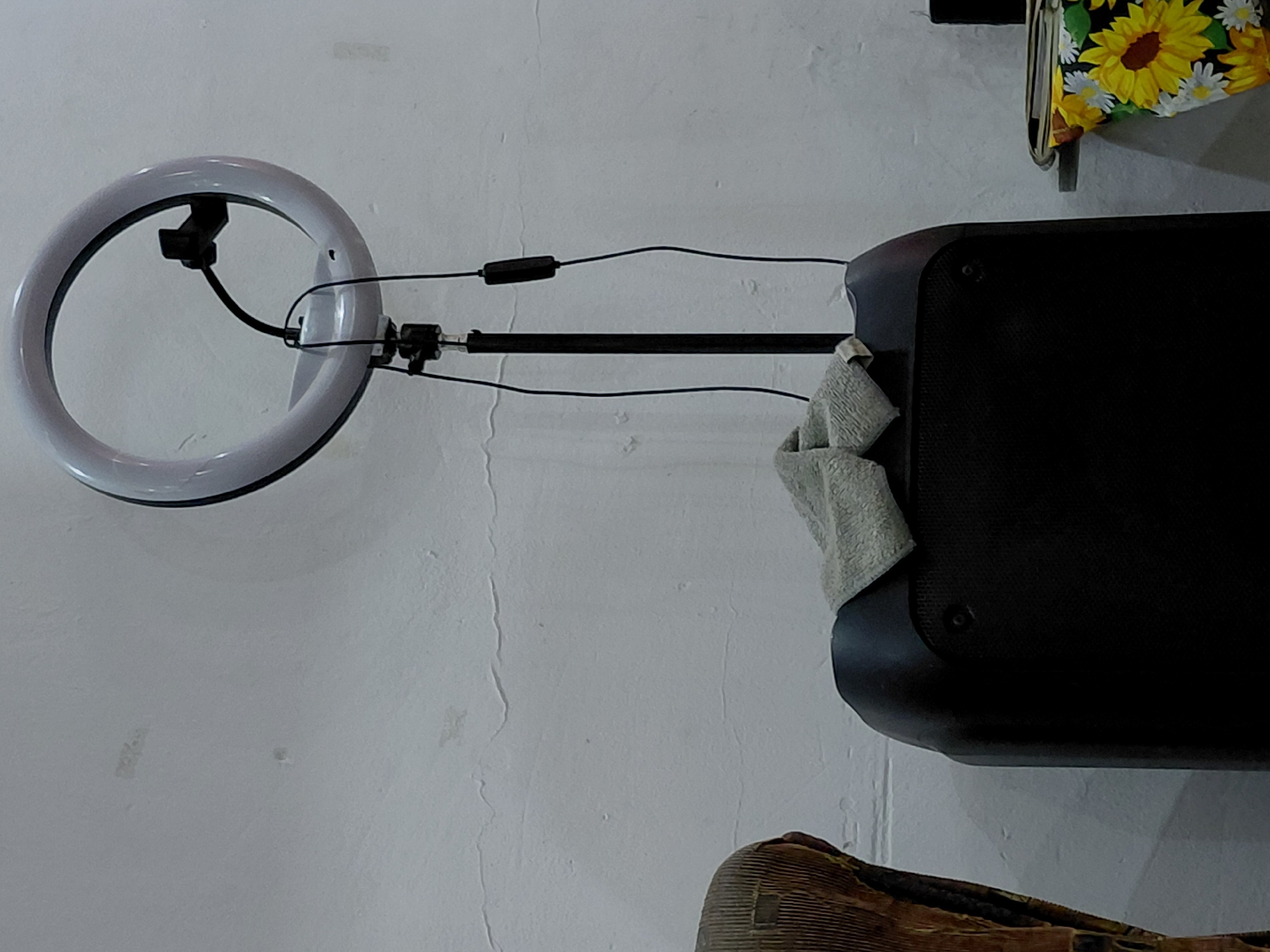 The image displays an indoor scene featuring a ring light, a black pole, and a black electronic device, likely a speaker or portable PA system, arranged against a white, textured wall. The ring light, unlit in the image, is positioned on the left and has a phone holder mounted in its center. Wires extend from the ring light, connecting to an inline control module and then along the black pole. This pole appears to be attached to, or part of, the black electronic device on the right side of the frame.
A light-colored, possibly white or light grey, towel is draped over the top right corner of the black electronic device. Above and slightly behind this device, a colorful bag with a distinctive yellow and white sunflower pattern is visible, hanging on a small hook or stand. In the bottom left corner, part of a brown, textured object, possibly a piece of furniture or another bag, is partially visible. The white wall in the background shows signs of wear, with visible cracks and uneven textures. There is no discernible activity taking place, nor is any text visible in the image. The setting appears to be a practical setup, potentially for recording or streaming, located in Santa Ana, Venezuela.