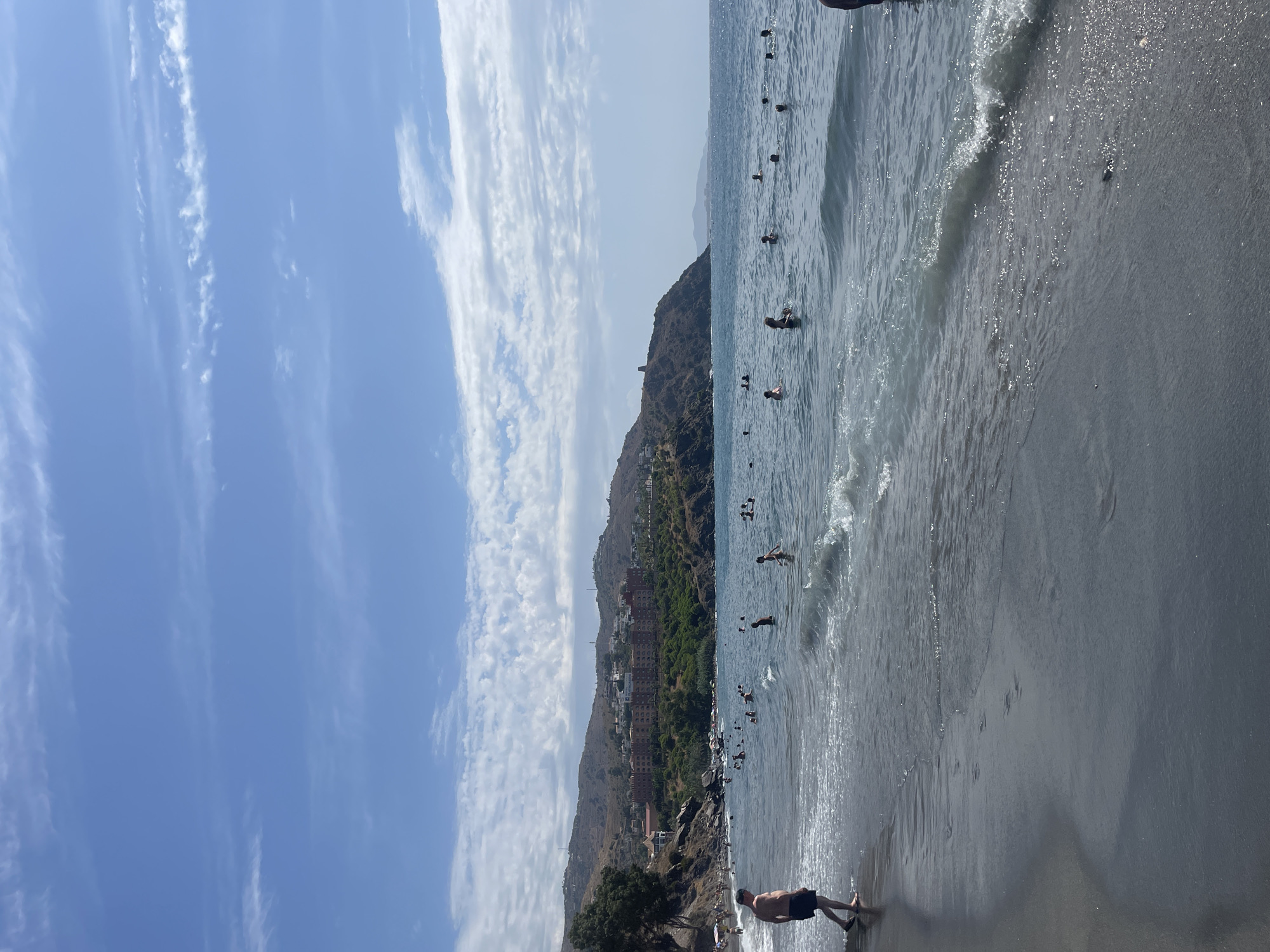 The image shows a sunny day at a beach in Almuñécar, Spain, with a hilly coastline in the background. The sea is calm with gentle waves lapping at the dark sand shore. Several people are seen swimming and wading in the water, appearing to enjoy a leisurely day. In the distance, a large hotel complex is visible nestled on the hillside, surrounded by lush greenery. The sky is blue with scattered white clouds, creating a picturesque summer scene. The foreground features the wet sand of the beach with some footprints visible.