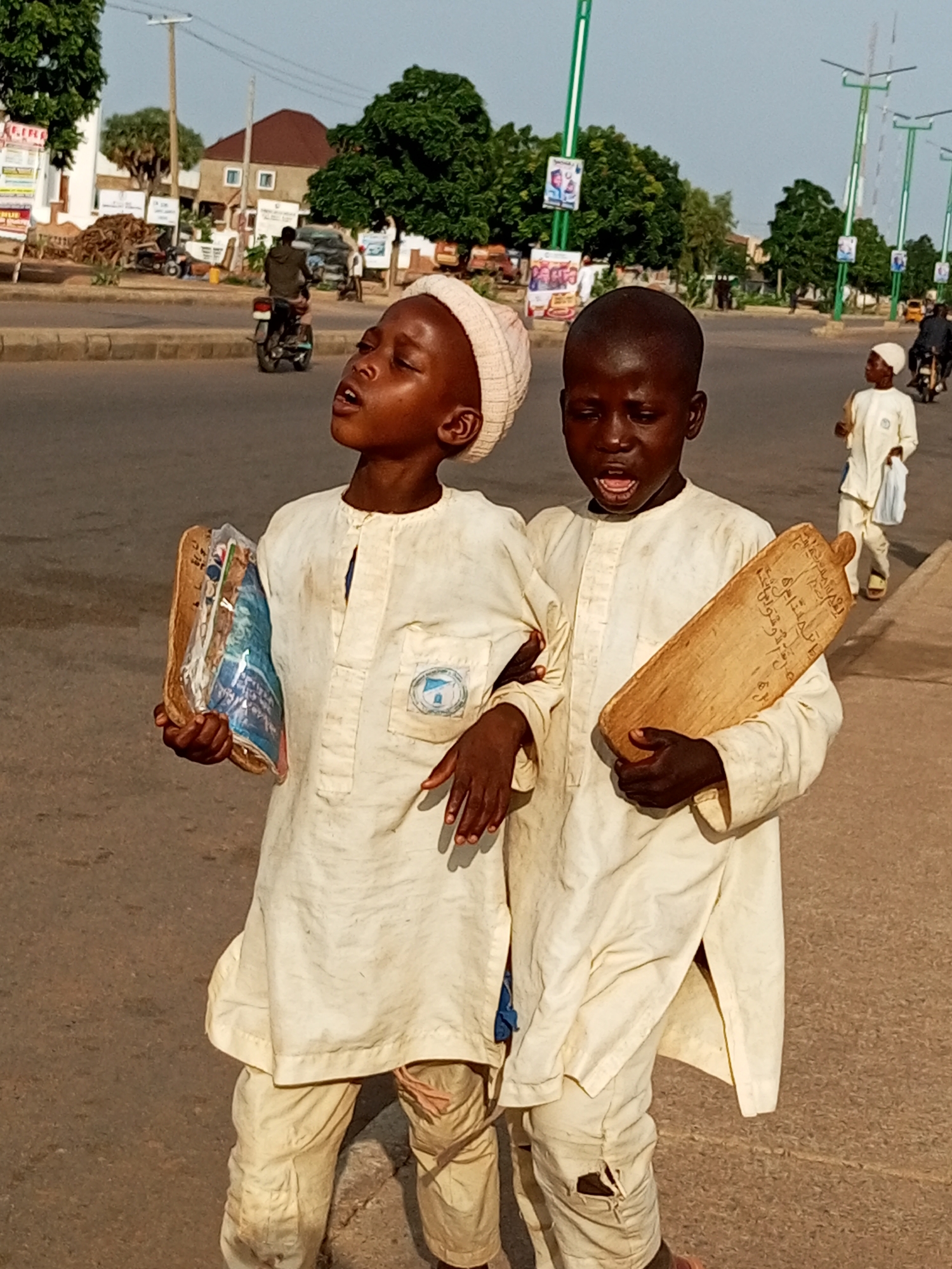 Two young boys, dressed in light-colored traditional clothing and head coverings, are walking side-by-side on a paved street in Tumfure, Nigeria. The boy on the left holds a stack of books wrapped in plastic, while the boy on the right carries a wooden slate with Arabic script. Both boys appear to be looking upwards and speaking, with their mouths open. In the background, other individuals on motorcycles and pedestrians are visible, as well as trees and buildings lining the street. The environment suggests a public urban area with ongoing activity.