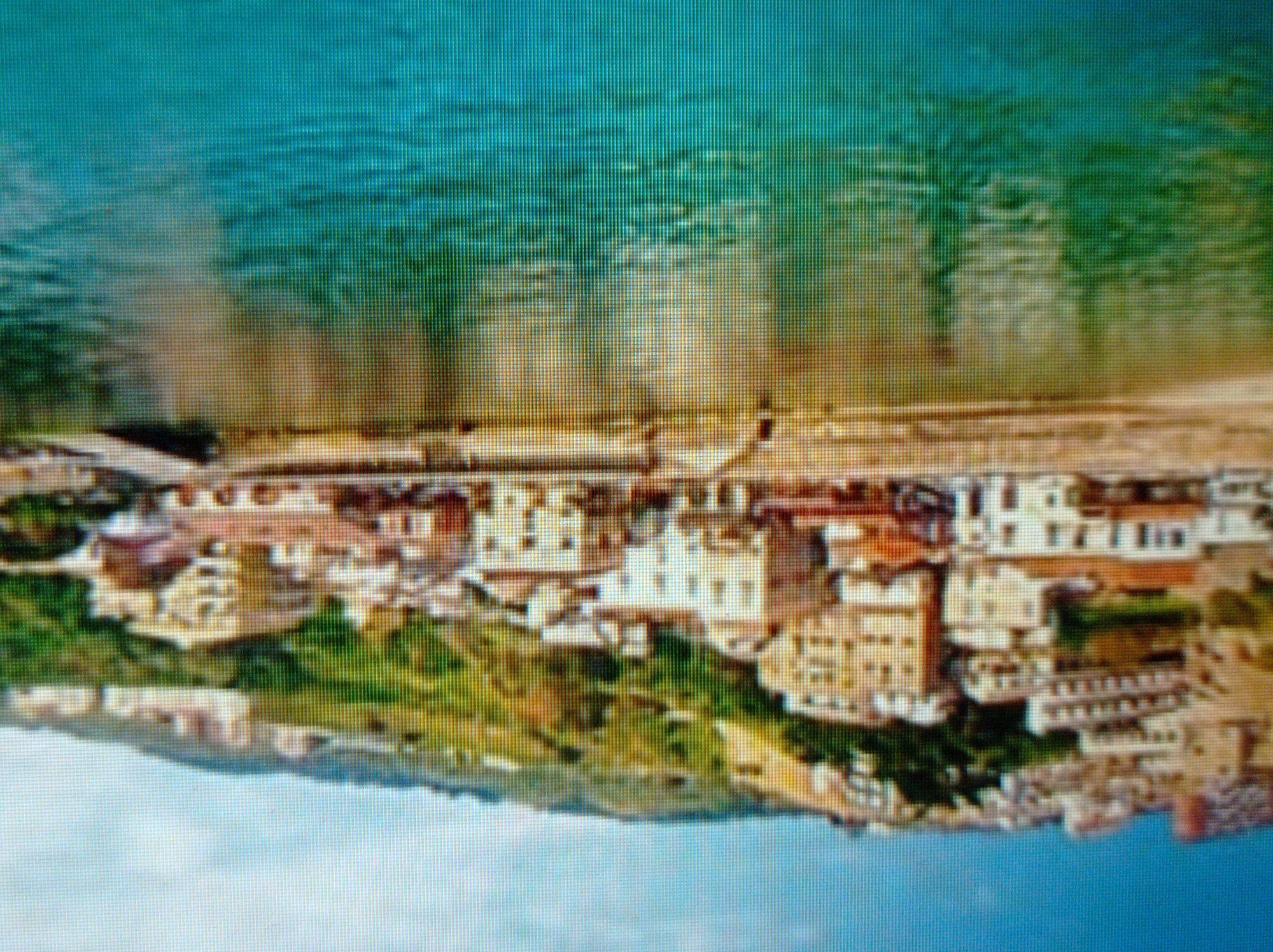 The image depicts a coastal city with buildings clustered on a hillside overlooking a body of water. The architecture appears Mediterranean, with light-colored structures, terracotta roofs, and balconies. Green vegetation covers the hillside behind the buildings. The water in the foreground is a vibrant turquoise and reflects the colors of the buildings and the sky. The image has a slightly blurred quality, suggesting it may have been taken from a distance or is a low-resolution photograph. There is no indication of this location being in Mukachevo, Ukraine, as Mukachevo is a landlocked city in western Ukraine and does not have a coastline.