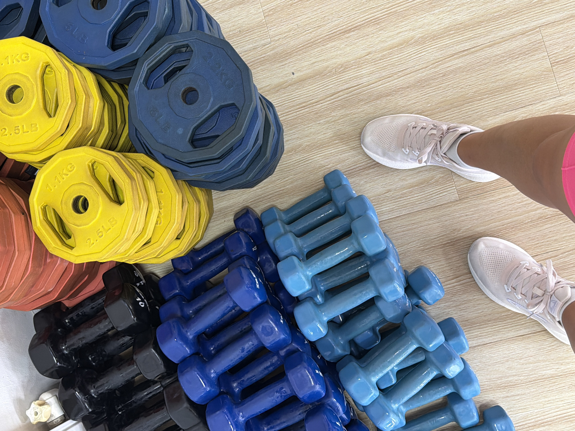 A high-angle shot shows a collection of weightlifting equipment and a person's legs and feet. The equipment is arranged on a wooden floor. There are stacks of round weight plates in yellow and blue, and several pairs of dumbbells. The dumbbells are in various shades of blue and black, some are heavier and some are lighter. The person's legs and feet are visible in the lower right corner of the image. They are wearing light pink athletic shoes and bright pink shorts. The background appears to be a wall with some white shelves. The scene is likely a gym or a home fitness area.