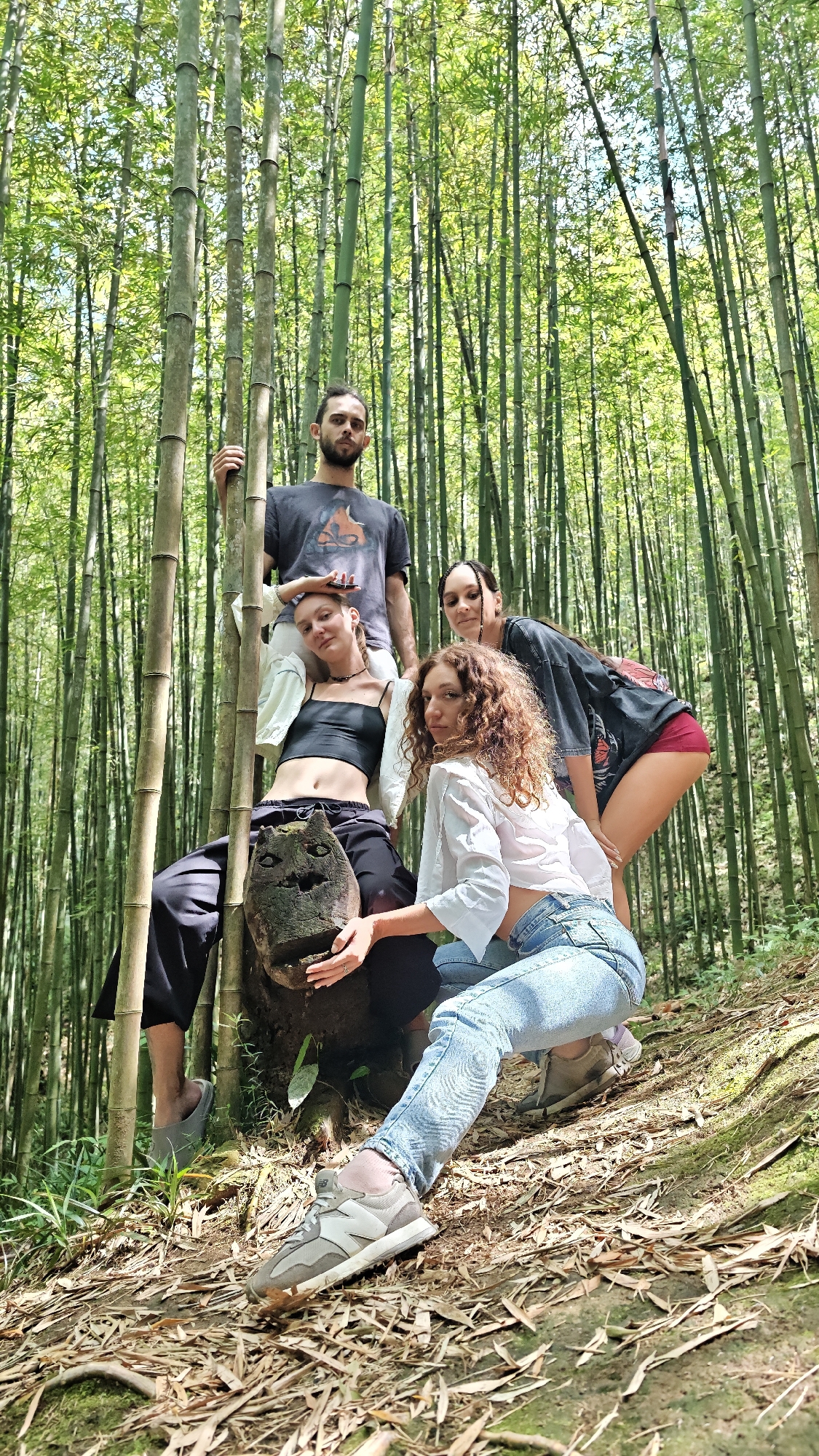 Four individuals are in a bamboo forest. A man in a dark gray t-shirt stands behind three seated or crouching women. A woman in a black crop top and loose black pants sits on a carved wooden object resembling a face. A woman with curly hair in a white crop top and jeans kneels to the right of the first woman. A woman in a black t-shirt and red shorts stands behind them. Bamboo stalks surround them. The ground is covered in fallen leaves and moss. The location is Ngã Ba Kim, Vietnam.