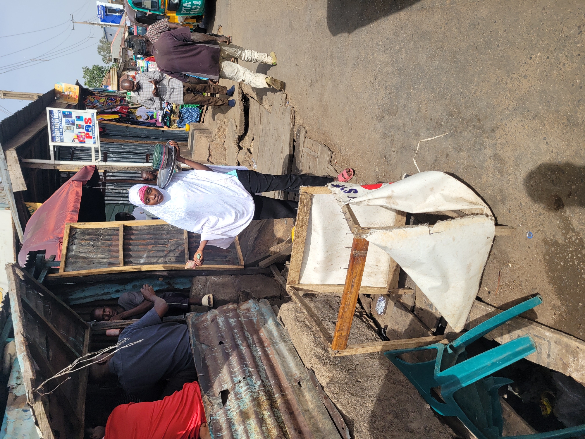 A street scene in Jos, Nigeria, depicts a woman wearing a white hijab and black pants holding a metal tray. She stands near a makeshift stall or shop constructed from corrugated metal and wood. Inside the stall, at least two men are visible. The stall also features a sign advertising "BUY PHONES ON EASYBUY AND IN CA HERE AND ACCESSORIES," with a phone number listed below. The stall also appears to be selling various items on display. A damaged, canvas-covered structure with wooden supports and a broken turquoise plastic chair are positioned on the ground nearby. Several individuals are walking along the street behind the stall, and a green tricycle or taxi is visible in the background. The overall environment appears to be a bustling marketplace or commercial area.