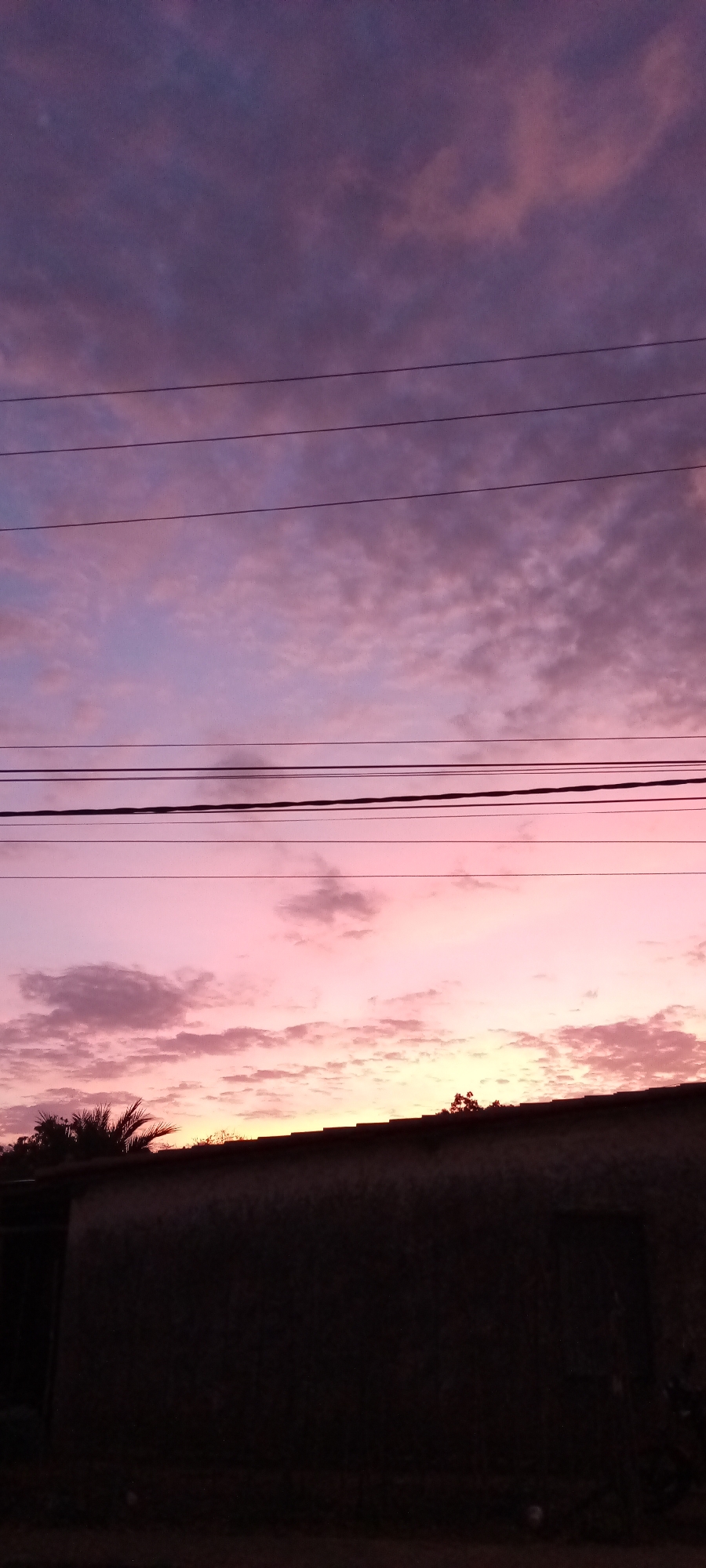 The image depicts a dusk or dawn sky with varying shades of pink and purple, punctuated by clouds. In the foreground, there is a dark, indistinct structure that appears to be a building with a visible roofline. Electrical wires stretch across the sky. The location is the city of Unknown, Unknown.