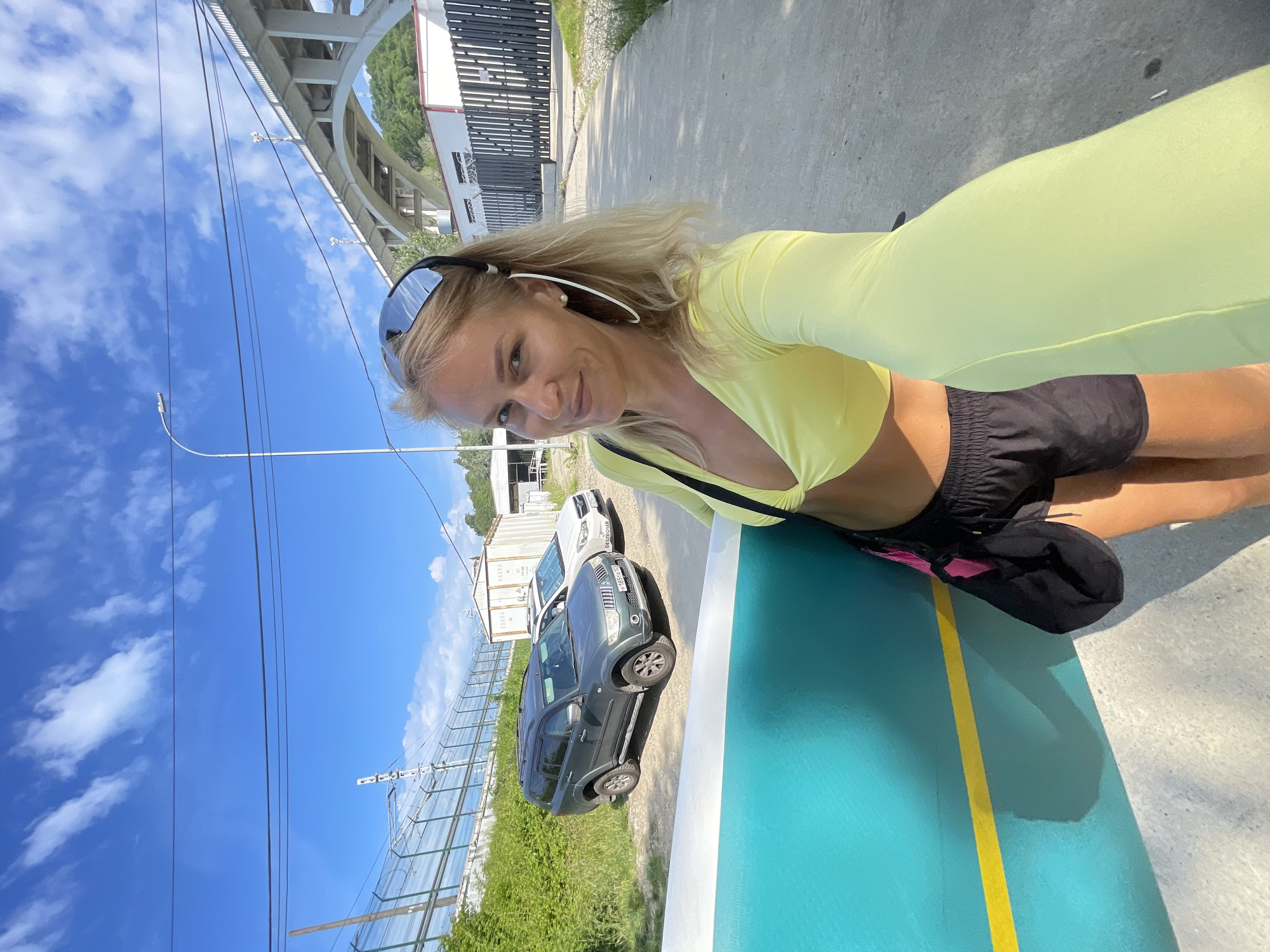 A blonde woman is taking a selfie while holding a teal and yellow surfboard outdoors on a sunny day. She is wearing a yellow long-sleeved crop top and black shorts. A black bag with pink accents hangs from her shoulder. In the background, a grey SUV and a white car are parked on a dirt road. Beyond the cars, there is a white shipping container and a dark fence. A concrete bridge with multiple arches spans the sky in the distance, and green hills are visible behind it. The scene appears to be in Sochi, Russia, possibly near a coastal area or waterway, given the presence of a surfboard and the bridge architecture.