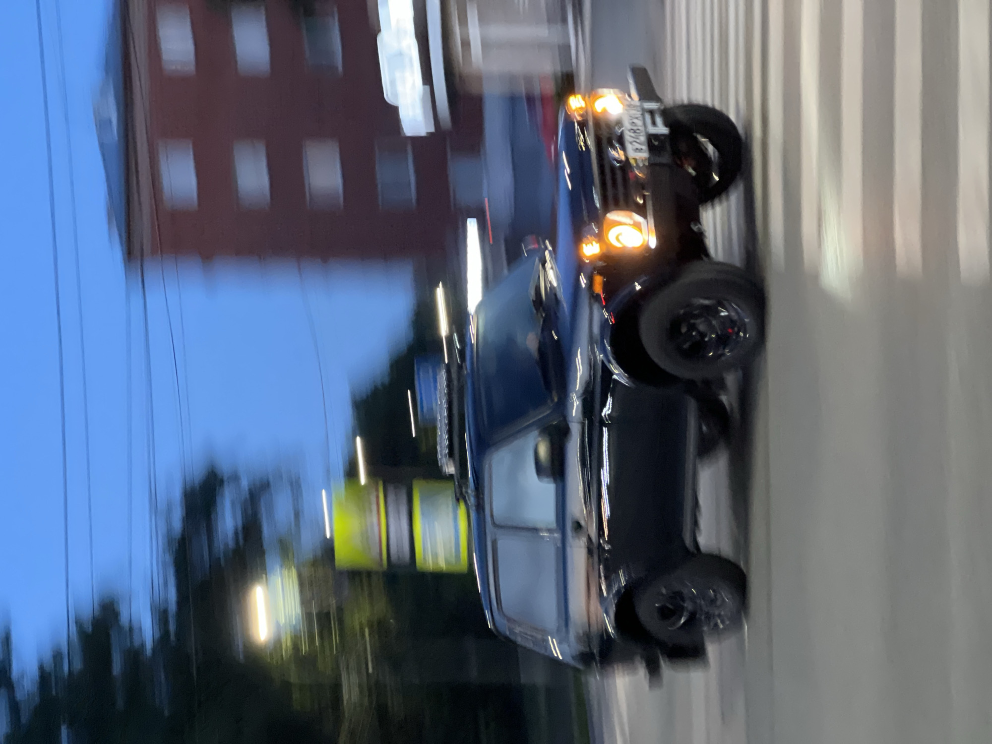 The image captures an urban scene in Izhevsk, Udmurtia, Russia, identified by the "18" region code on the vehicle's license plate. A black two-door Lada Niva SUV, likely a 3-door variant, is centrally positioned, moving from left to right across a pedestrian crossing marked with white stripes on an asphalt road. Its headlights and amber turn signals are illuminated. The vehicle is equipped with a roof rack and features black-painted wheels. The Russian license plate "B248PX 18" is clearly visible on the front bumper. A blurred silhouette may be present in the driver's seat; no other individuals are clearly discernible.
The background shows significant horizontal motion blur, characteristic of a panning photographic technique or the photographer being in motion. To the left, blurred dark green foliage, streetlights with bright horizontal streaks of light, and an indistinct, illuminated yellow-green sign are present. In the mid-ground, a multi-story, reddish-brown brick residential building with multiple rectangular windows is heavily blurred. To the right of this building, another structure with a brightly illuminated, blurred white sign, possibly indicating a commercial establishment, is visible. Overhead, multiple power lines traverse a blue twilight sky.