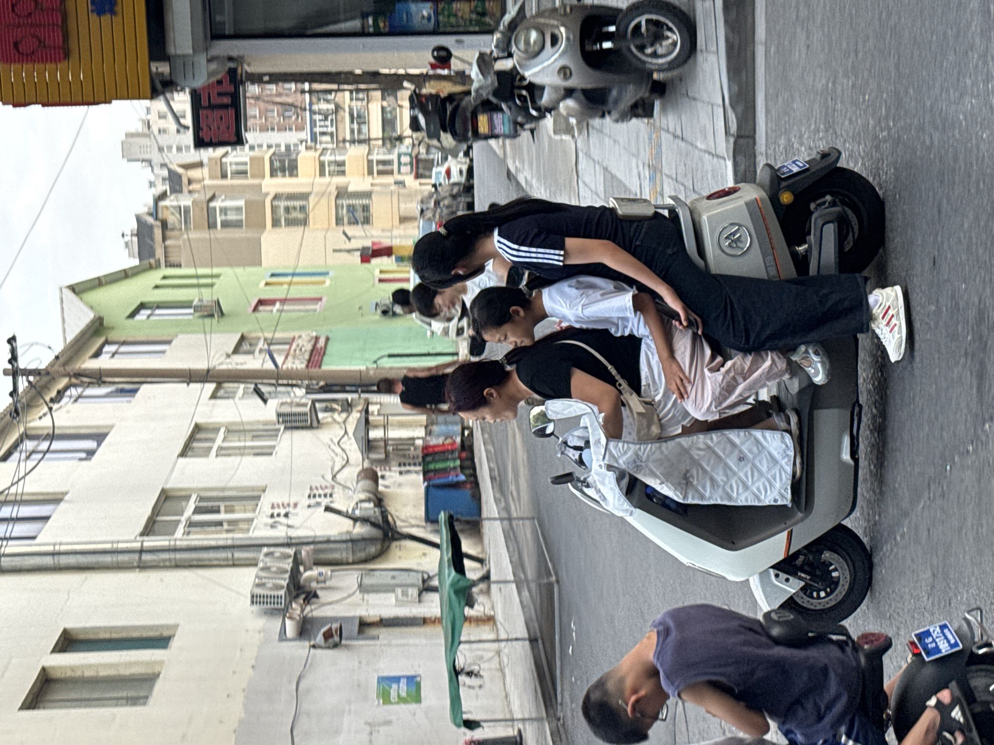 In Guta, China, on a street with mixed-use buildings, a family of four rides on a white electric scooter. A woman in a black shirt and white shorts is driving, with a younger girl in a white shirt and pink pants seated between her and another girl in a black Adidas-striped shirt and black pants. A young boy in a purple sleeveless shirt is in front of them on the same scooter. In the background, other scooters and cars are visible, along with buildings of various colors, including white, green, and beige. The scene appears to be in the daytime under an overcast sky.