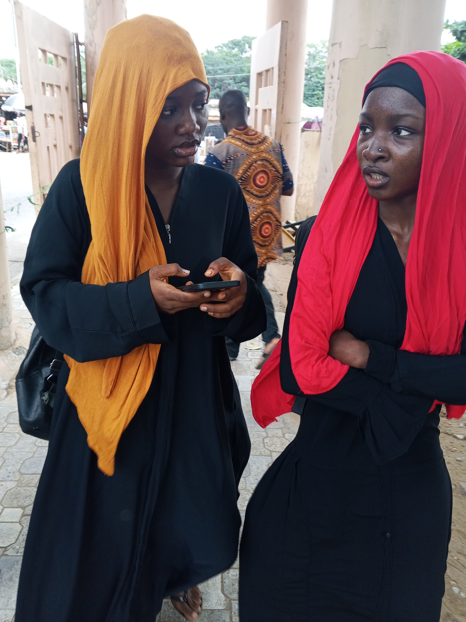 Two young women are standing on a paved outdoor area in Lokoja, Nigeria. The woman on the left is wearing a black abaya and an orange headscarf. She is looking at her phone, which she holds in her hands. The woman on the right is wearing a black abaya and a red headscarf. She is looking to her right with her arms crossed. In the background, there are some market stalls, a person wearing a patterned shirt, and some trees. The environment appears to be a public space, possibly near an entrance to a building or a market.