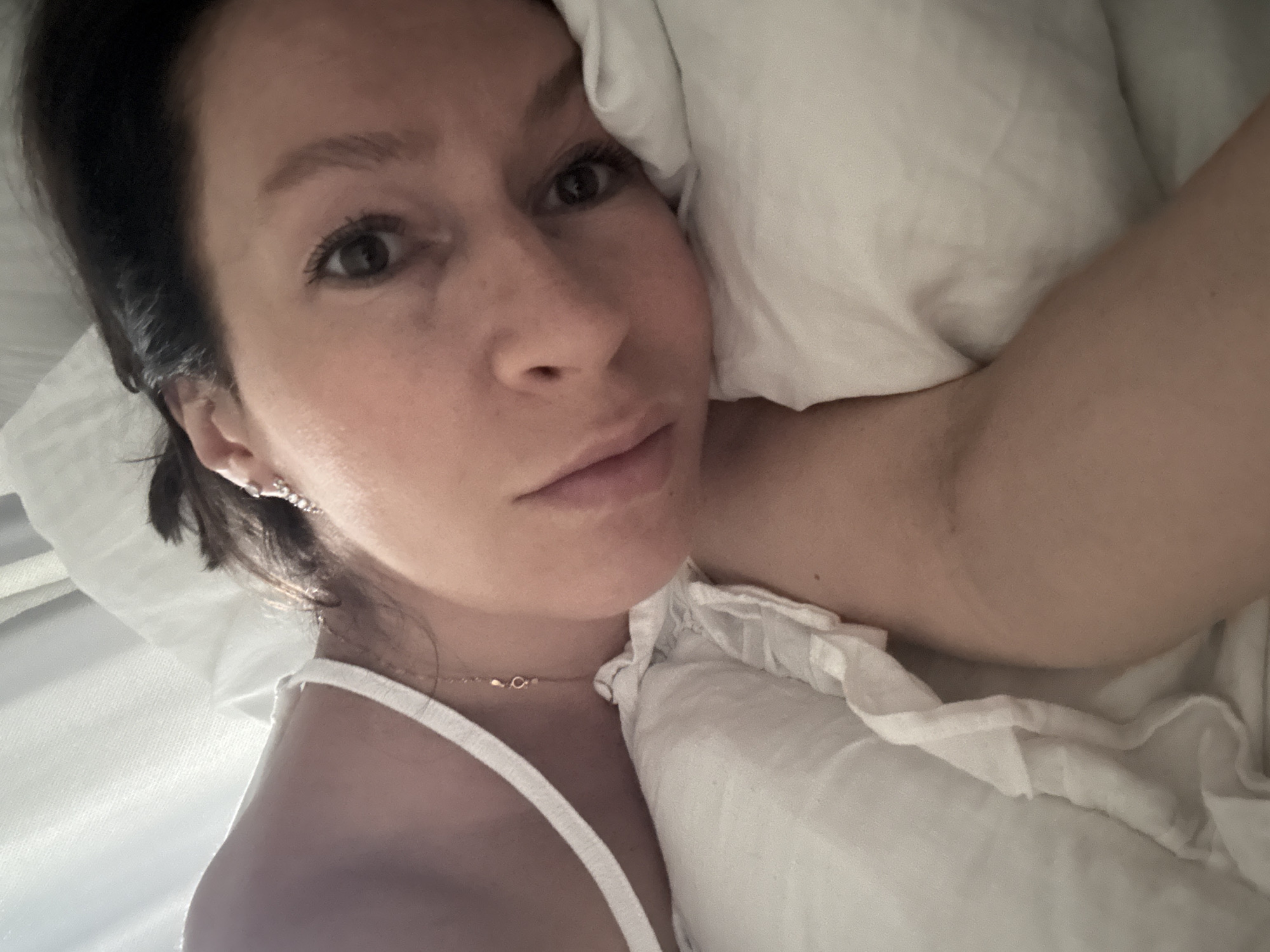 A woman is lying in bed. She is on a white pillow and is wearing a white top and a gold necklace. She has brown hair and brown eyes and is looking at the camera. She is wearing earrings. The location is unknown and cannot be determined to be Chaikovsky, Russia, from this image.