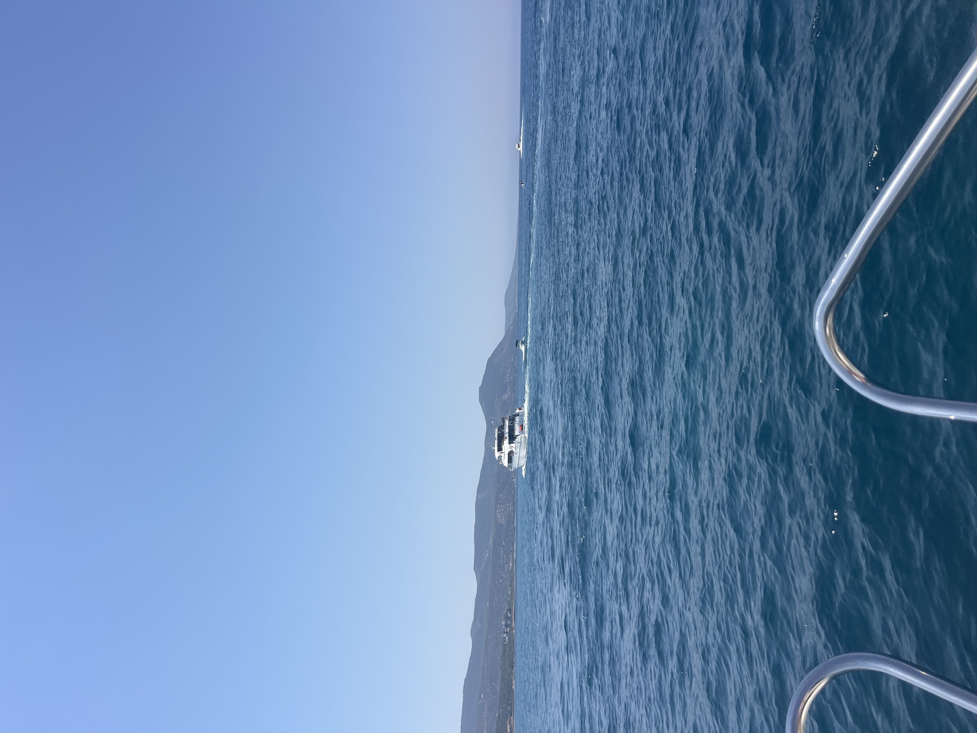 An offshore view from the front of a boat in Polis Chrysochous, Cyprus, shows a large white passenger boat moving away from the camera. The boat has multiple decks and a dark roof. Behind the passenger boat, a small, dark object is visible on the water, possibly a jet ski or another small craft. Further in the distance, another white boat is moving at speed, leaving a wake. The background consists of a low, green, mountainous coastline under a clear blue sky. The water is a deep blue with small waves. The foreground of the image shows the polished metal railings of the boat the photo is taken from.