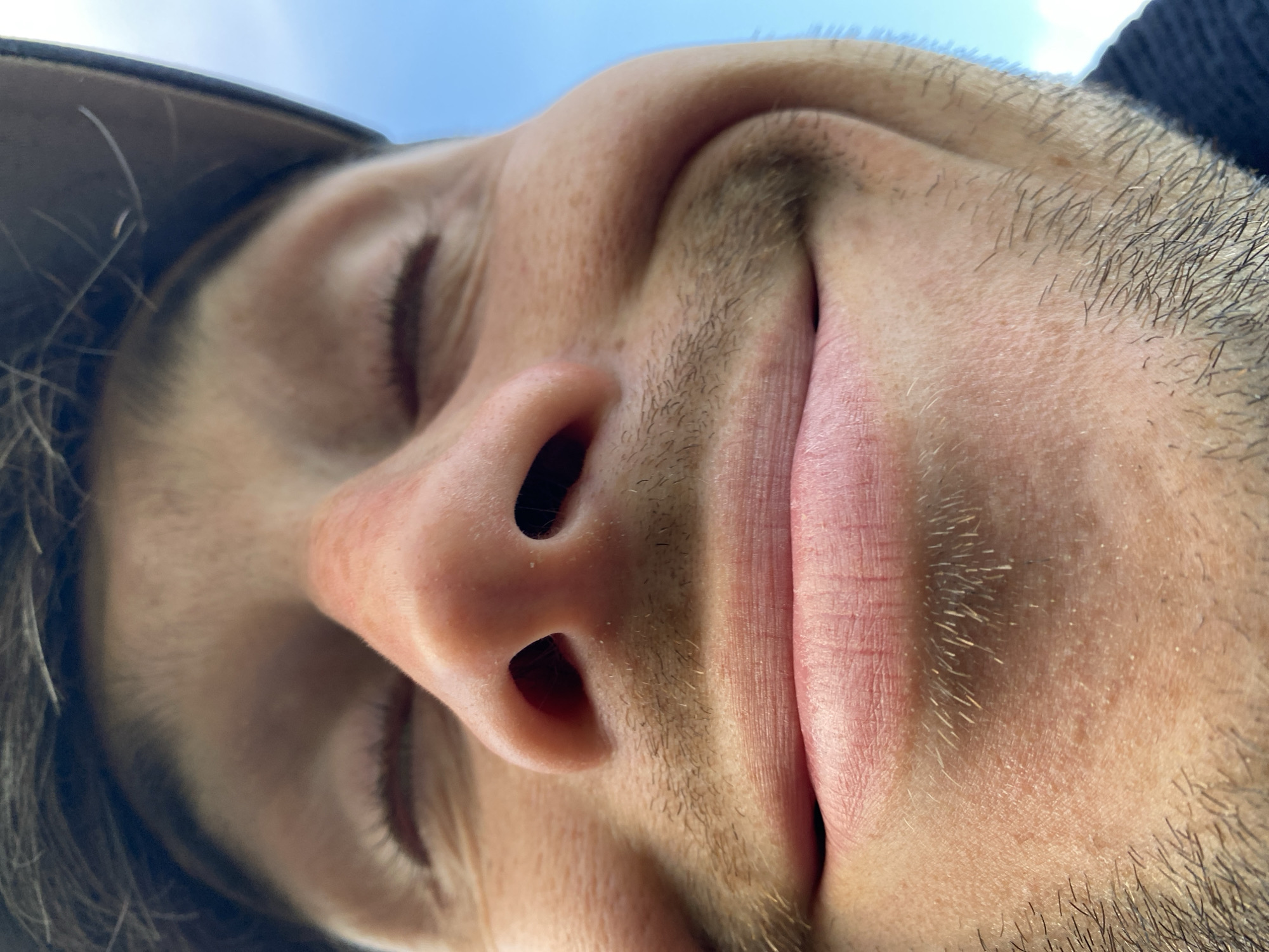 This is a close-up, low-angle shot of a person's face, tilted upside down. The focus is on their nose and mouth, with their eyes closed. The person appears to be outdoors, as indicated by the bright sky visible in the upper portion of the image. The lighting suggests it is daytime. The individual has light-colored skin and is wearing a dark cap, a portion of which is visible on the left side of the frame. Short, dark hairs are visible on their upper lip and chin, indicating some facial hair. The overall mood is relaxed, possibly even peaceful, given the closed eyes and slight upward curve of the lips suggesting a subtle smile. The image was taken in Oktyabr'skii, Russia. There is no visible text in the media file.
