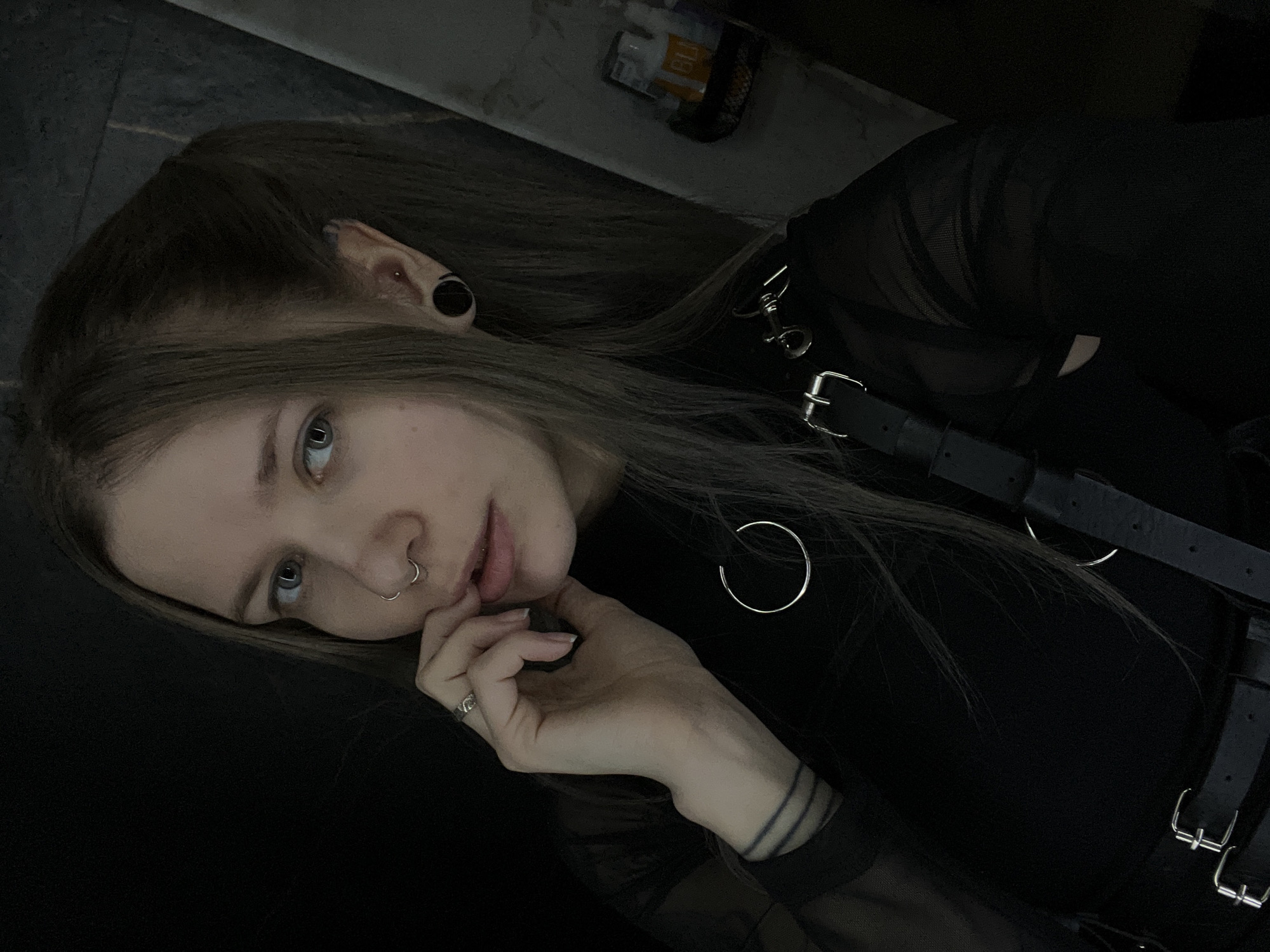 A young person with light skin and shoulder-length brown hair is lying on a dark surface. The person has piercings in their nose and ear. They are wearing a black top, leather straps and buckles. The person's left hand is up near their mouth. It is not possible to determine the location as Khimki, Russia based on the image.