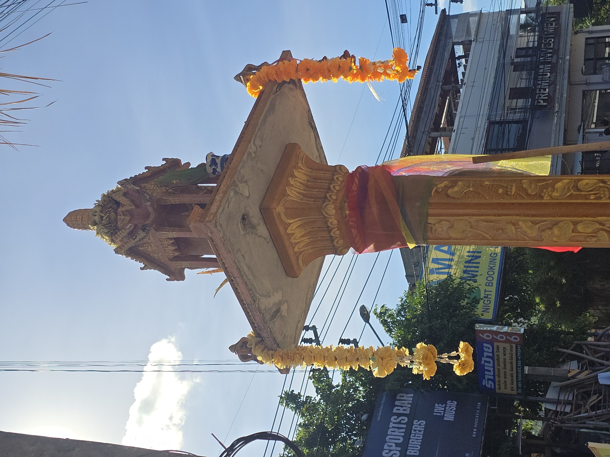 An outdoor, eye-level, medium shot captures a spirit house decorated with garlands, set against a bright blue sky with scattered white clouds. The spirit house, made of concrete and adorned with gold accents, stands on a decorative pillar draped with colorful fabric. Yellow and orange flower garlands hang from the spirit house. In the background, there are power lines, trees, streetlights, and buildings, including one with a sign that reads "SPORTS BAR." Another building has a sign with "PREMIUM INVESTMENT" and other businesses are visible. The overall scene suggests an urban environment in a tropical or subtropical region.