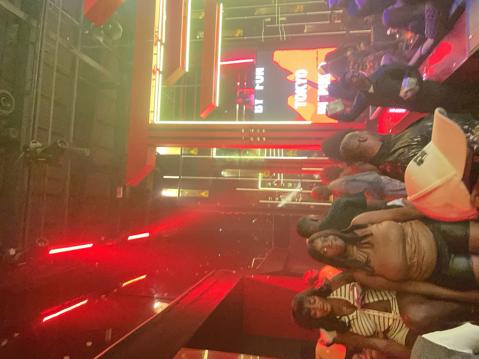The image depicts an indoor scene at a nightclub or event space, possibly in Abuja, Nigeria, with a focus on the ambient lighting and the presence of patrons. Red and yellow lights create a vibrant and energetic atmosphere, with prominent red lighting on the left side of the frame and a large illuminated sign featuring the words "TOKYO" and "INSPIRE" in the background. A DJ or performer is visible behind a console in front of the "TOKYO INSPIRE" sign. Several individuals are in the foreground and mid-ground, some looking towards the camera and others engaged in their own activities. The overall environment suggests a lively social gathering.