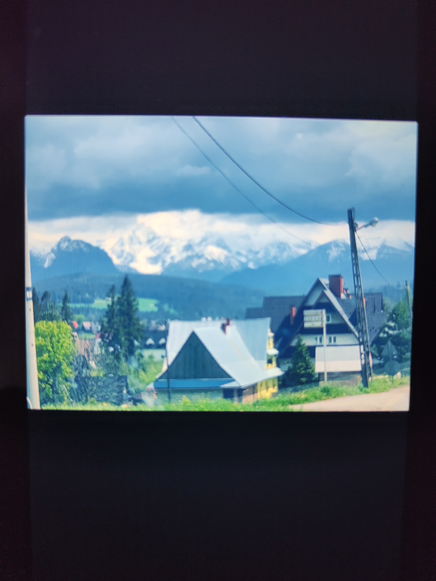 This media file depicts a landscape scene featuring houses, trees, and snow-capped mountains under a cloudy sky. In the foreground, there are two houses. The house on the left has a light-colored roof and yellow walls, with some green trees and foliage in front of it. The house on the right is larger, with a dark roof, a red chimney, and a light-colored facade. A tall, dark utility pole with a streetlamp is situated to the right of this house.
In the middle ground, there are more trees, including evergreen trees, and the roofs of other buildings can be seen, suggesting a village or town. The background is dominated by a range of mountains, with their peaks covered in snow, under a sky filled with dramatic, dark clouds. The overall impression is of a somewhat overcast day in a mountainous region, possibly in spring or autumn, given the presence of green vegetation alongside the snow. There is no visible text in the image.