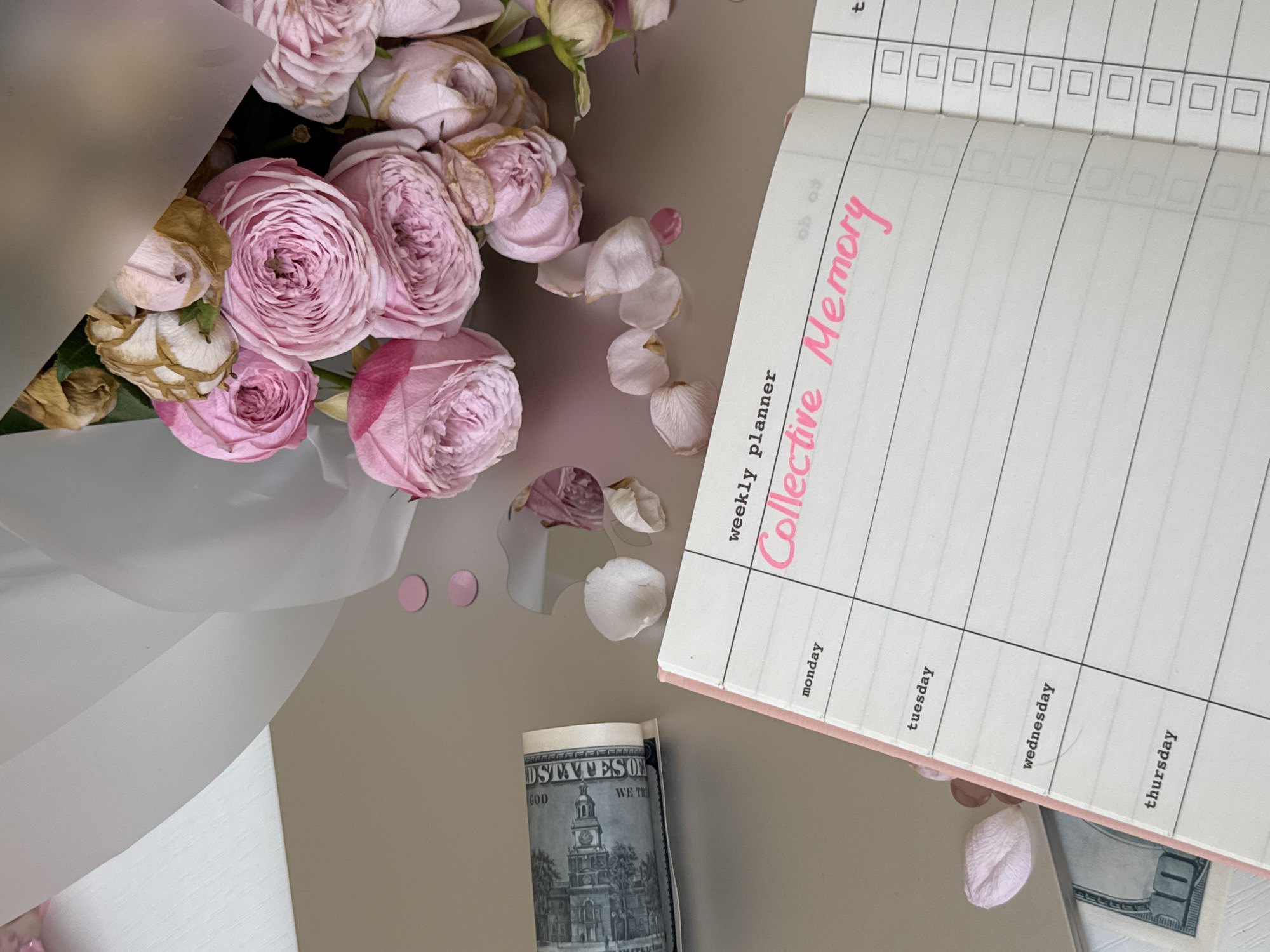 The image depicts a close-up, overhead view of a desk surface. A bouquet of pink roses, some of which are wilted, is prominently placed on the right side of the frame. In the center, an open weekly planner with a pink cover is visible. The planner has lines for each day of the week, with "weekly planner" written at the top and "Collective Memory" handwritten in bright pink marker across the main section. To the left of the planner, a rolled-up US dollar bill is present. Scattered around the objects are pink confetti circles and a few fallen rose petals. The background consists of a light-colored wall on the left and a slightly out-of-focus area of the desk surface. There are no indications of a specific location in Yekaterinburg, Russia. The scene appears to be staged for a still life composition, with no people or discernible activity.