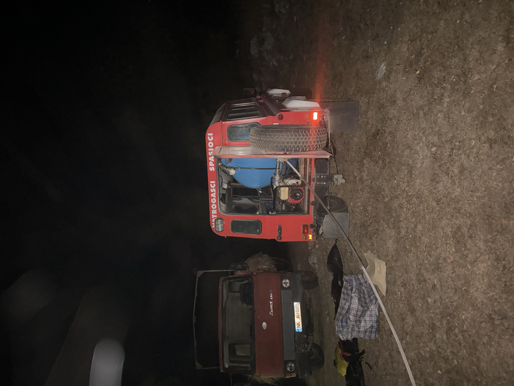 The image shows two vehicles, a red truck and a red fire truck, parked on a dirt or gravel surface at night. The red truck on the left appears to be older, with a visible license plate that reads "NK AU885". It is loaded with some sort of hay or dry vegetation in its cargo bed. The red fire truck on the right is a four-door vehicle with a large blue tank mounted on the back, along with some firefighting equipment. The text "VATROGASCI SPASIOCI" is visible on the side of the fire truck, indicating it is for firefighters and rescuers. Several bags and a rolled-up cloth are on the ground in front of the vehicles. The background is dark and appears to be a natural or rural environment, possibly with some rocky terrain and vegetation visible in the distance. The scene is illuminated by artificial light, likely from the vehicles or external sources, creating shadows and highlights. There are no people visible in the image. The location is identified as Bijele Poljane, Montenegro.