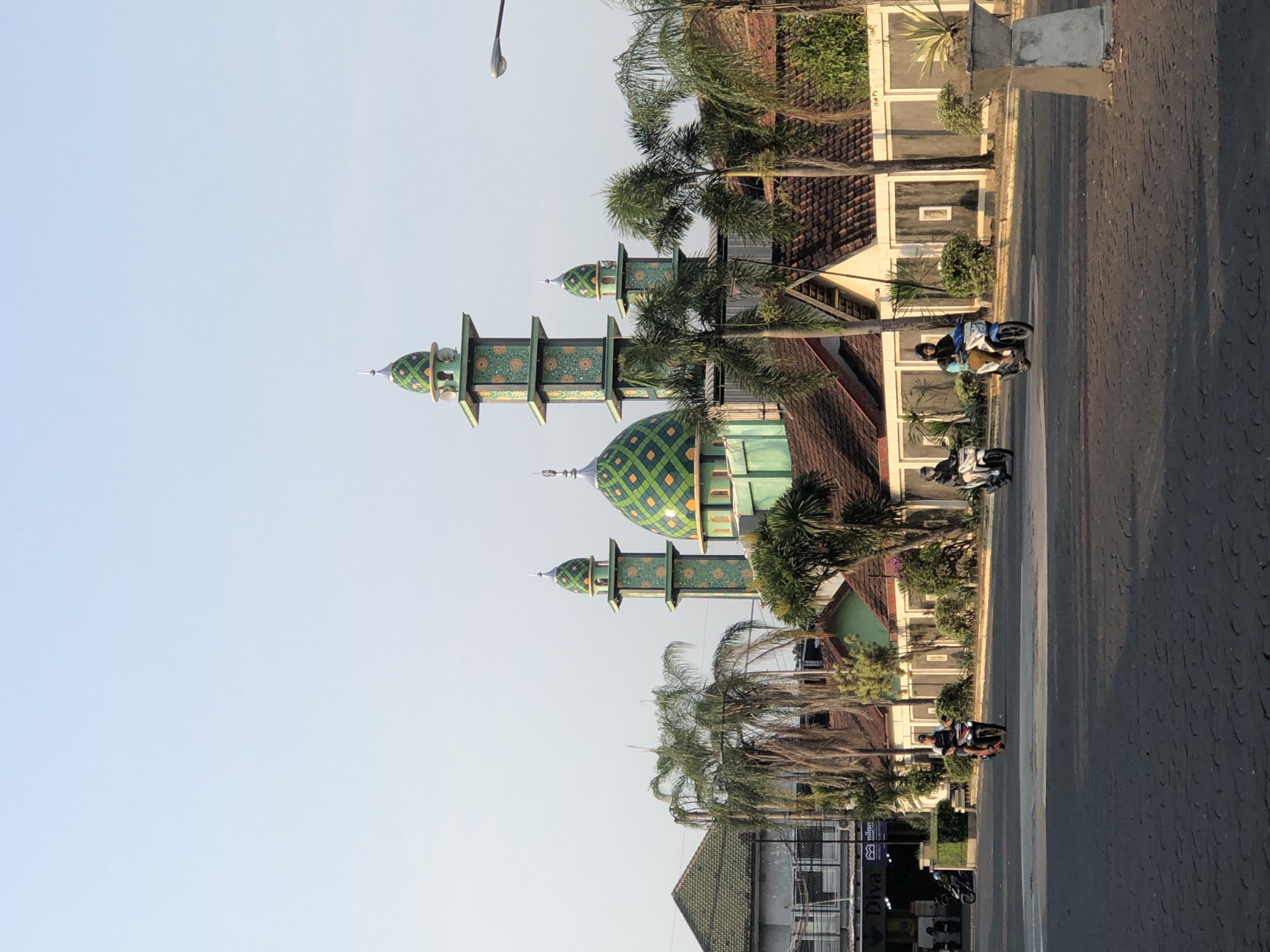 In Boro, Indonesia, a mosque with a vibrant green and yellow patterned dome and multiple minarets stands prominently against a clear, pale blue sky. The mosque's architecture features ornate green towers adorned with geometric patterns. In front of the mosque, palm trees line a road where several individuals are riding motorcycles. The road is paved with dark interlocking blocks, and there are buildings with terracotta roofs visible behind the mosque, some with white facades. A streetlamp is visible on the right side of the frame.