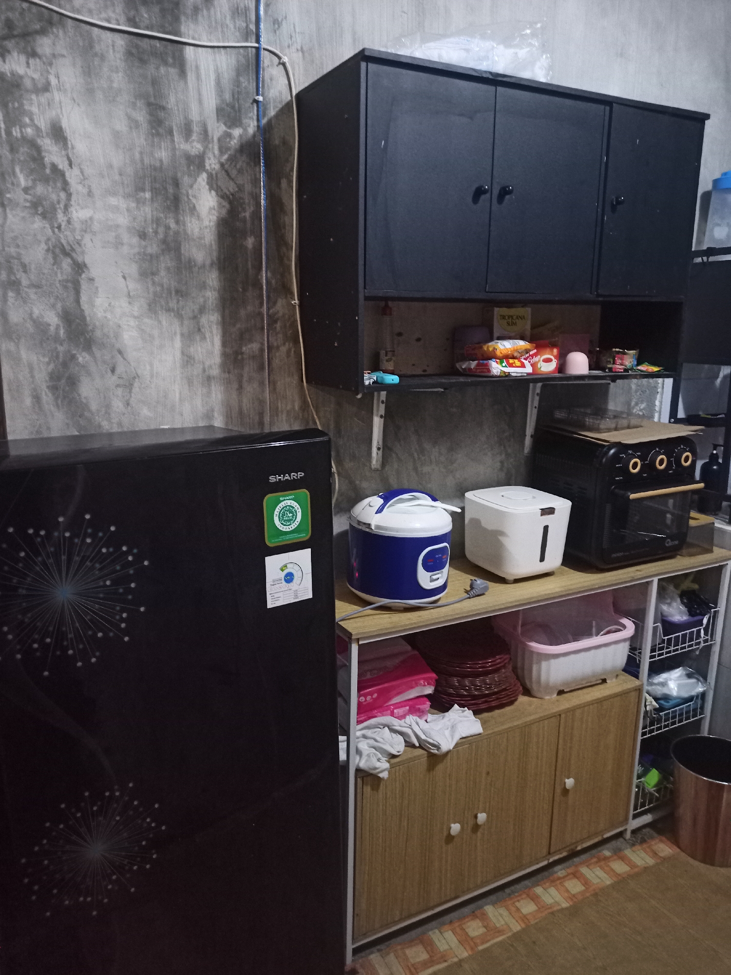 The image captures a corner of a kitchen or storage area in a residence located in Randuagung, Indonesia. The scene is dimly lit, with a rough, textured concrete wall forming the backdrop. On the left, a black Sharp refrigerator with a decorative dandelion motif is visible. To the right of the refrigerator, a two-tiered shelving unit is present. The upper shelf holds a black cabinet with three doors, beneath which are several packaged snacks and a small pink object. On the lower shelf of the unit, a blue and white rice cooker, a white square appliance, and a black air fryer oven are arranged. Below this shelf, a wooden cabinet with two doors sits atop a metal-framed shelf unit. This shelf unit contains various household items, including pink storage containers, stacks of red plates, and wire baskets filled with more items. A metallic trash can is positioned to the far right. The floor is covered with a patterned mat. A blue cord runs vertically along the concrete wall, secured by clips.