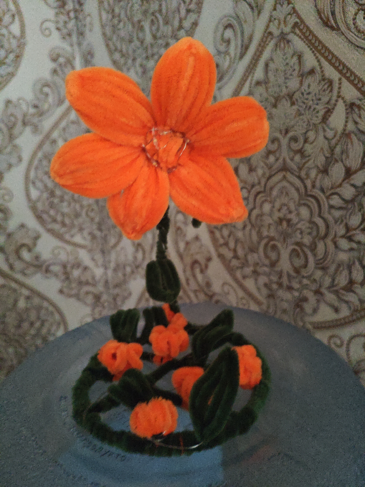 An orange chenille flower with five petals sits atop a thin green stem. The flower has a shiny, slightly reflective center. Below the flower, a ring of dark green chenille stems encircles several small orange chenille pumpkins and green leaves. This arrangement rests on a gray, circular surface. The background is a wall with a repeating ornate pattern in shades of brown and cream. The image appears to be taken indoors, likely on a table, and there is no indication of the location being Krasnaya Polyana, Russia.