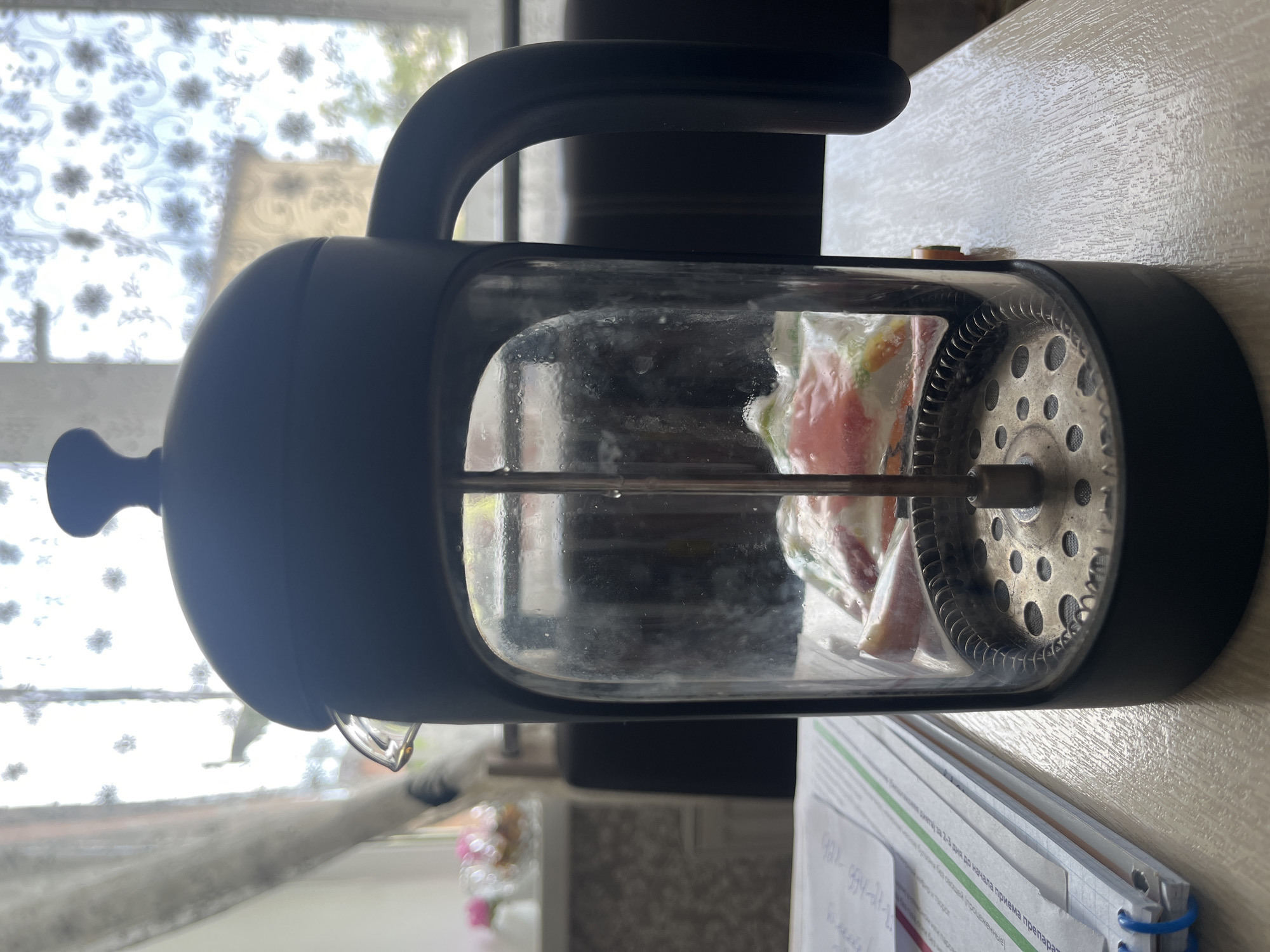 The image shows a French press coffee maker with a black handle and frame, and a glass carafe. Inside the carafe, there is a sealed plastic bag containing what appears to be fruit pieces, possibly for making infused water or a fruit tea. The French press is positioned on a light-colored, textured surface, likely a table or countertop. In the background, a window with white floral patterned curtains is visible, allowing diffused daylight to illuminate the scene. A stack of papers or a notebook lies to the right of the French press, with some handwritten text partially visible. The overall lighting suggests it is daytime. The scene appears to be indoors, in a domestic setting. No people are visible in the image. The text on the papers is in Cyrillic script and is not clearly legible.