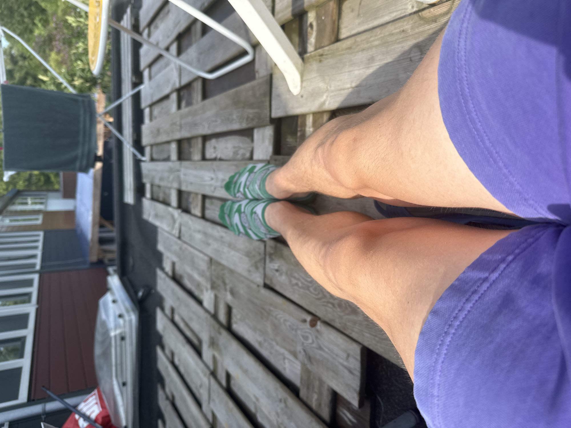 A person is lying on a wooden deck, with their legs extended towards the camera. The person is wearing purple shorts and green and white striped socks. The wooden deck is made of weathered planks and appears to be on a rooftop. In the background, there is a building with a dark red and blue facade. There is also a clothes drying rack with a dark green towel hanging on it, and a small yellow table with white chairs. The scene suggests a relaxed outdoor setting, possibly a balcony or terrace. The location is identified as Lelystad, Netherlands.
