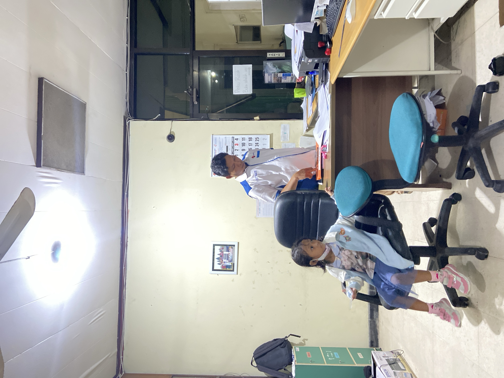 A rotated, indoor shot features a man in a white and blue shirt sitting at a desk and interacting with a young girl in a blue skirt and pink shoes. The scene appears to be an office with a calendar on the wall, a closed door with a sign "Pintu Rusak, Buka sebelah sini" (Broken Door, Open This Side) on the upper part, a monitor on the desk, and various office supplies. A black backpack and printer are on the floor. The location is inferred to be Surabaya, Indonesia, based on the inscription on the door sign.