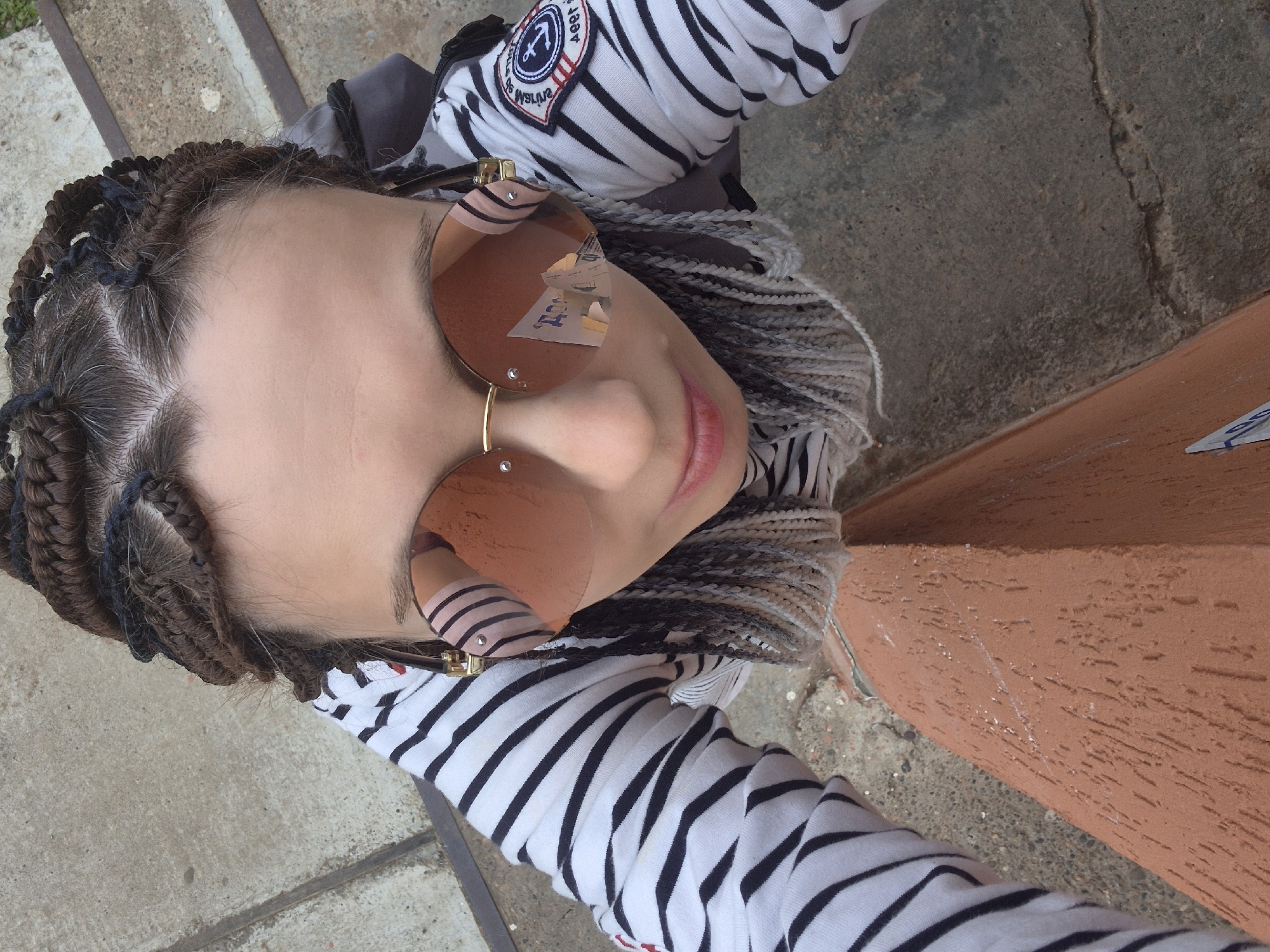 This media file is a close-up, high-angle, outdoor selfie of a young person with braided hair. The subject is wearing a black and white striped long-sleeved shirt and large, round, rose-tinted sunglasses. The braids are a mix of brown, black, and gray. The subject's face is tilted upwards, and their mouth is slightly open, with pink lipstick visible. The reflection in the sunglasses shows what appears to be a receipt or a piece of paper with some text and images.
The setting appears to be outdoors, with a concrete ground visible in the foreground and background, suggesting an urban or park-like environment. A textured, reddish-brown surface is visible on the right side of the image, possibly a wall or a railing. The lighting suggests it is daytime, likely sunny, given the strong reflection in the sunglasses. The overall mood is casual and confident. The location context is Chaikovsky, Russia, though this is not visually evident in the image itself. The shirt has a patch on the left sleeve with an anchor and some text, but the text is not clearly legible.