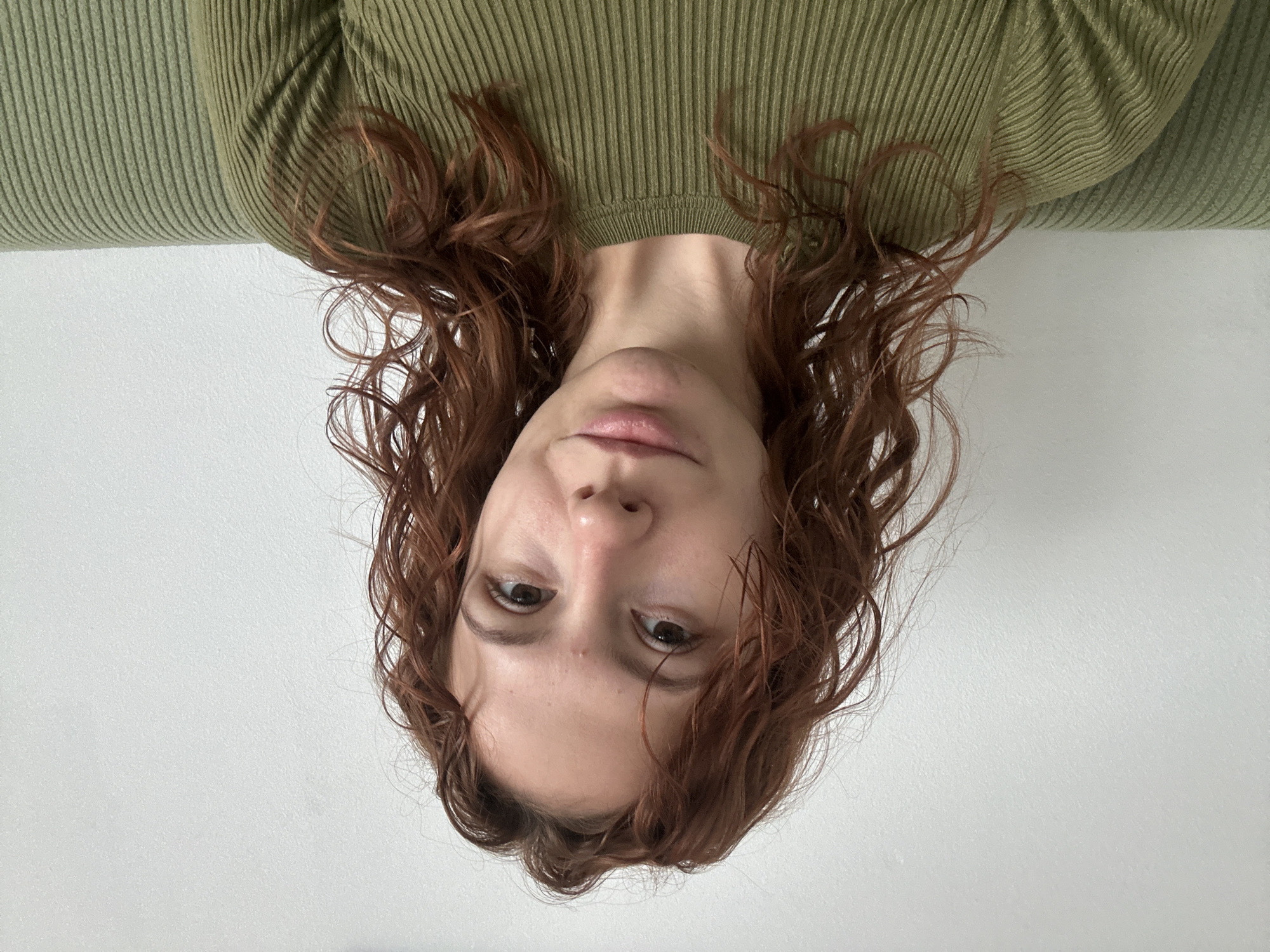 A woman with curly, reddish-brown hair is positioned upside-down in front of a white wall. She is wearing a ribbed, olive-green top. The image offers a close-up view of her face, revealing features like her nose, lips, and eyes. The background is plain and white, suggesting a room with a neutral backdrop. The location is unknown; there is no indication that the image was taken in Novi Sad, Serbia.