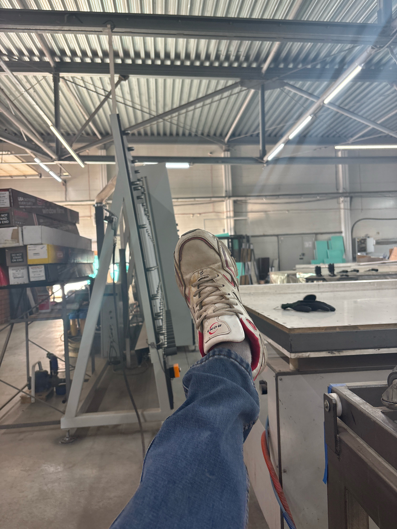 The image depicts an indoor industrial setting, possibly a workshop or factory. In the foreground, a person's right leg, clad in blue denim jeans and a white and red athletic shoe, is raised and positioned in front of the camera. The shoe exhibits signs of wear and has a visible brand name "BONA" on it.
The background reveals a large, open space with a corrugated metal ceiling supported by exposed metal beams and trusses. Fluorescent lighting fixtures are mounted on the ceiling. Several pieces of industrial equipment are visible. To the left, a tall, grey machine with a vertical panel and an angled support frame is present. Stacked boxes are also visible on a metal shelving unit. In the center and right of the frame, a white work surface or table is prominent, with a pair of black gloves resting on it. Other machinery and materials are distributed throughout the space. The overall environment suggests an active manufacturing or processing facility. There is no indication of the location being specifically Barnaul, Russia.