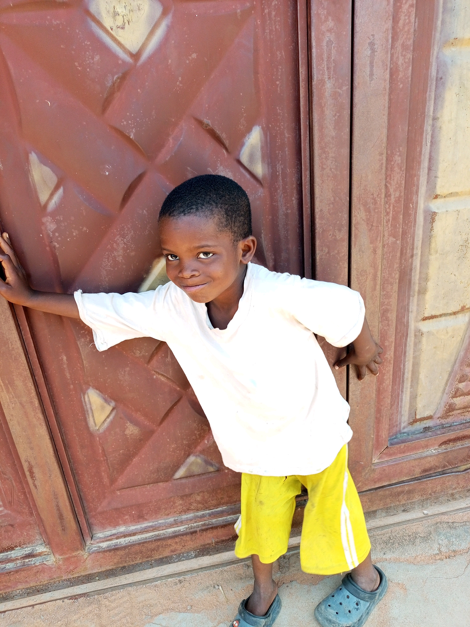 A young Black child, appearing to be around 6-8 years old, stands with their left arm resting on a large, reddish-brown metal door. The child is wearing a plain white t-shirt and bright yellow shorts with white stripes. They are looking directly at the camera with a slight smile and have short, dark hair. The door has a diamond-shaped pattern embossed on its surface and appears to be part of an exterior wall. The ground beneath the child is dusty or sandy. The overall impression is of an outdoor setting, possibly in a residential area of Tempure, Nigeria, where the child is posing for a photograph.