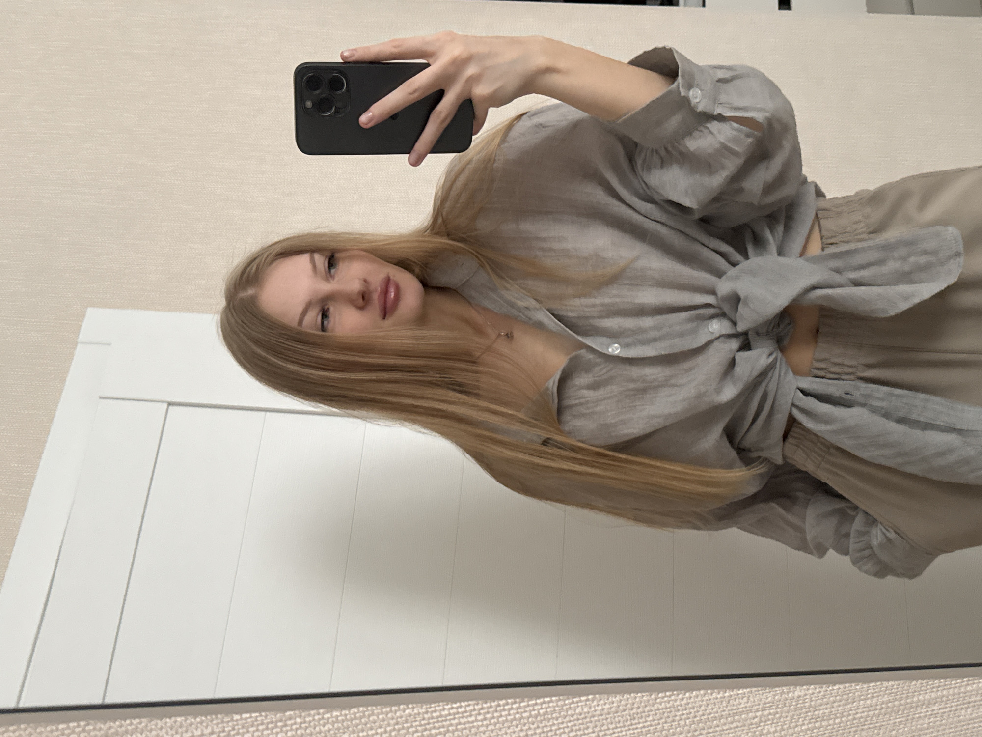 A young woman with long blonde hair is taking a mirror selfie in what appears to be a bedroom. She is wearing a grey oversized linen shirt tied at the waist, revealing a sliver of her midriff, and light-colored pants. She is holding a black smartphone in her right hand, angled to capture her reflection. The background shows a white paneled door or closet to her left and a textured beige wall to her right. The overall impression is casual and domestic. This image is not taken in the city of Perm, Russia; there are no identifying markers in the image to suggest this location.