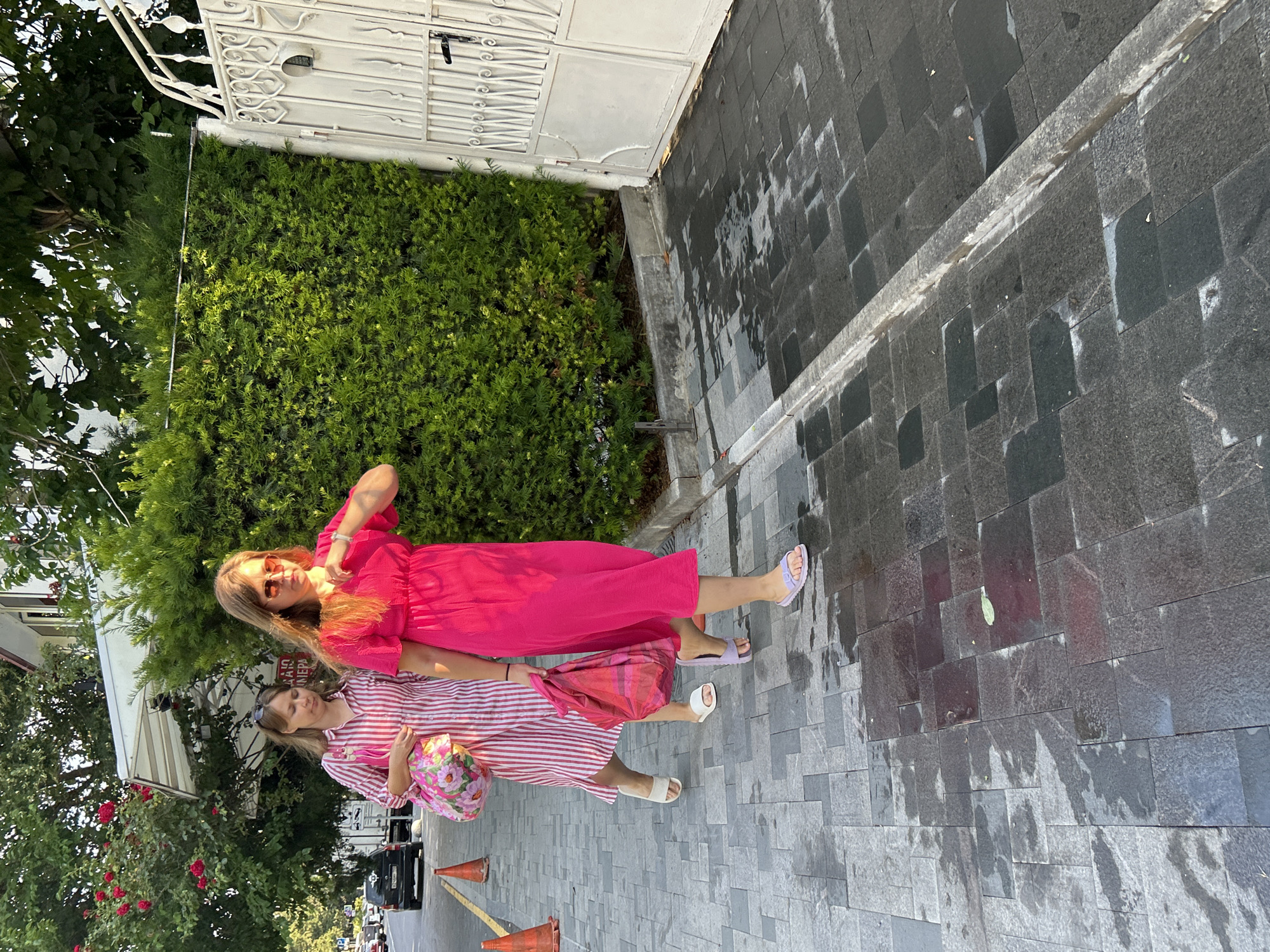 Two women are walking on a wet paved sidewalk in front of a green hedge and a white metal gate. The woman on the left wears a pink and white striped dress and carries a floral patterned bag. The woman on the right wears a bright pink dress and carries a pink tote bag. Both women are wearing sunglasses and sandals. In the background, a car is parked on the street, and orange traffic cones are visible. The scene appears to be in Gelendzhik, Russia, indicated by the presence of red roses and a typical Mediterranean architectural style in the background building.