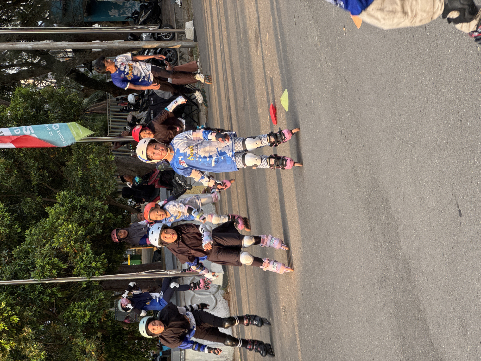 In Banda Aceh, Indonesia, a group of children are rollerblading on an asphalt path under a tree-lined environment during daylight hours. The main subjects are four young girls wearing helmets and protective gear, engaged in rollerblading activities. The girl in the foreground, on the right, wears a blue and white long-sleeved shirt with checkered pants and pink rollerblades. To her left, another girl in a black hijab and a long-sleeved black top with blue accents also wears pink rollerblades. Further to the left, a third girl with a black hijab and a blue and black long-sleeved shirt is on rollerblades. Behind them, a fourth girl in a red helmet and a patterned long-sleeved shirt is also rollerblading. In the background, several adults and older children are present, some on motorcycles and others observing. A red and green flag is visible on a pole, and trees with dense foliage provide shade. The setting appears to be a public outdoor space, possibly a park or recreational area.