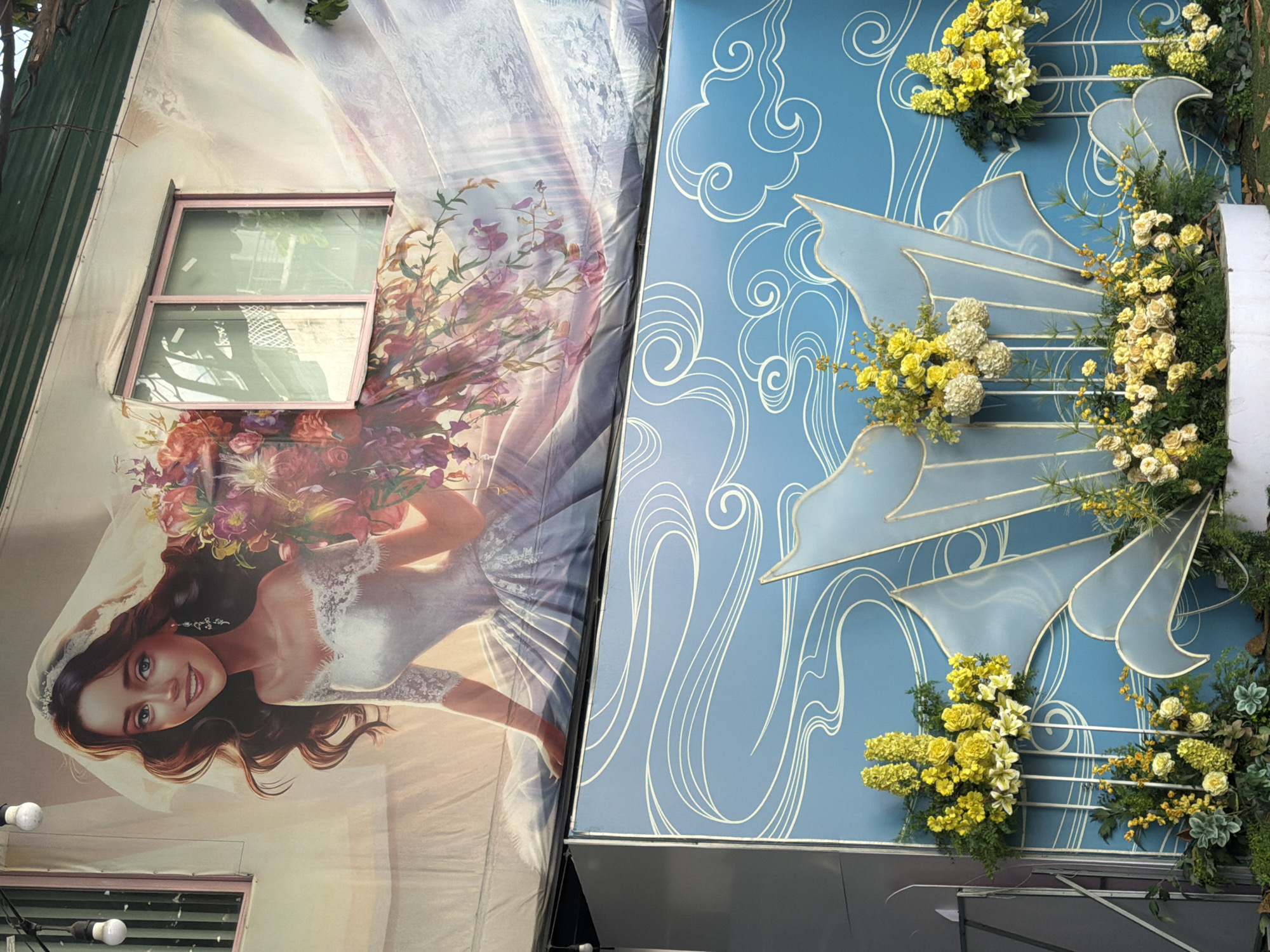 A mural depicting a bride in a wedding dress is painted on the exterior wall of a building. The bride has long brown hair, a veil, and holds a bouquet of flowers. The mural extends over a window with pink trim. Below the mural, a blue wall with white cloud-like patterns serves as a backdrop for a floral arrangement. The arrangement consists of yellow and white flowers, with greenery, placed on white stands. The scene appears to be outdoors, with visible greenery and a suggestion of a roofline at the top of the image.