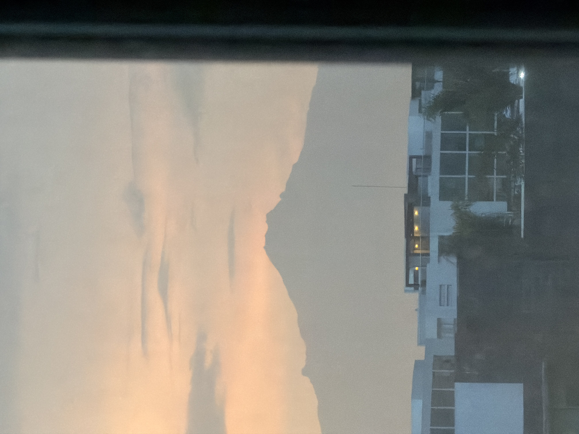 The image, taken from a window, shows a view of the urban landscape of San Andrés Cholula, Mexico. On the left side, there is a large, light-colored mountain range, likely reflecting the light of the setting sun. To the right, a modern, multi-story building with a grid-like pattern of windows is visible. The building's architecture suggests a contemporary design with possible commercial or residential use. Trees or foliage partially obscure some parts of the building. The image has a muted, slightly hazy quality.