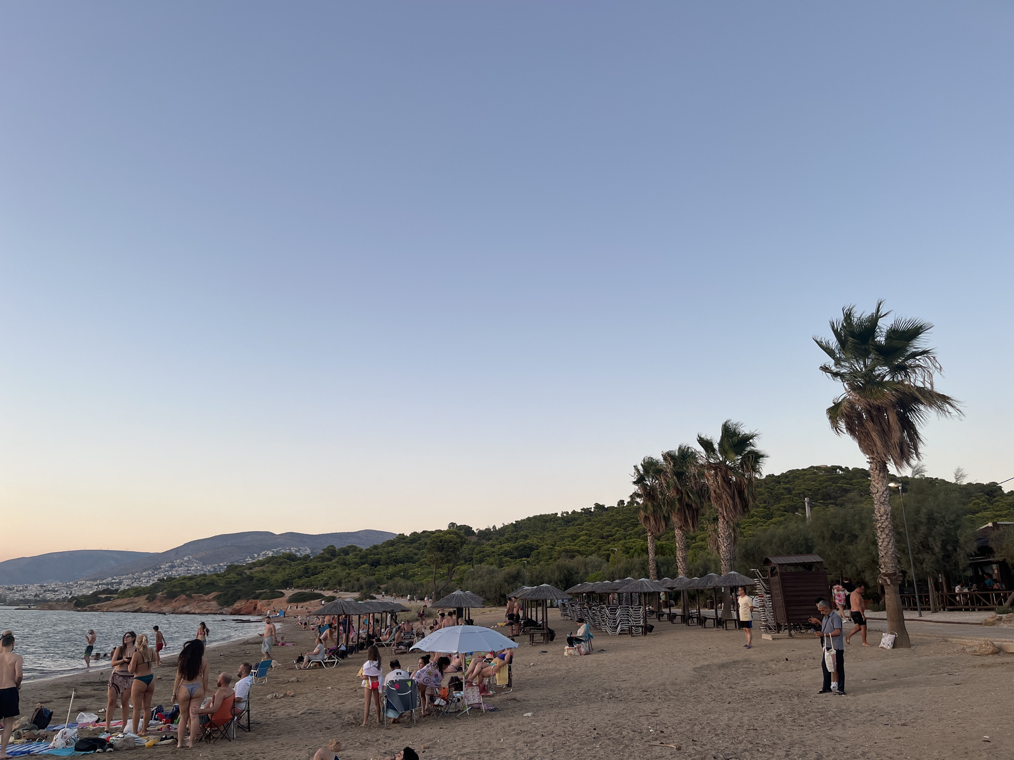 The image depicts a crowded beach in the Vouliagmeni Municipal Unit, Greece, during what appears to be late afternoon or early evening. The sky is a clear, pale blue, transitioning to a lighter hue near the horizon. In the background, rolling hills covered in dense green vegetation and scattered houses form a backdrop to the scene. The beach itself is sandy, with a line of umbrellas and lounge chairs set up for visitors. Several palm trees are scattered along the coastline, adding to the tropical feel.
Numerous people are visible on the beach, engaging in various activities. Some are standing and conversing, others are sitting on chairs or lounging on the sand. Several individuals are in or near the water, suggesting swimming or wading. Beach towels and bags are spread on the sand. A small structure resembling a changing room or kiosk is situated on the right side of the image, near a wooden deck and more umbrellas. The overall impression is one of a vibrant, active beach scene with a mix of relaxation and social interaction.