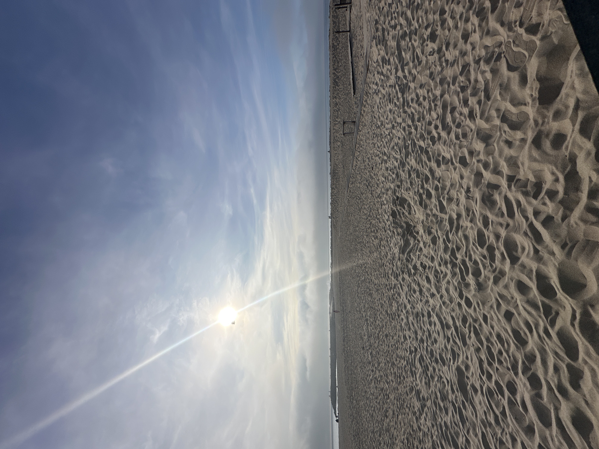 The image shows a beach in Espinho, Portugal. The scene is divided between a sandy beach on the right and a sky with the sun on the left. The sand is textured with wave-like patterns. The sky is mostly blue with some white clouds, and the sun is shining brightly near the center-left, creating a bright glare. A few people are seen walking in the distance along the beach.