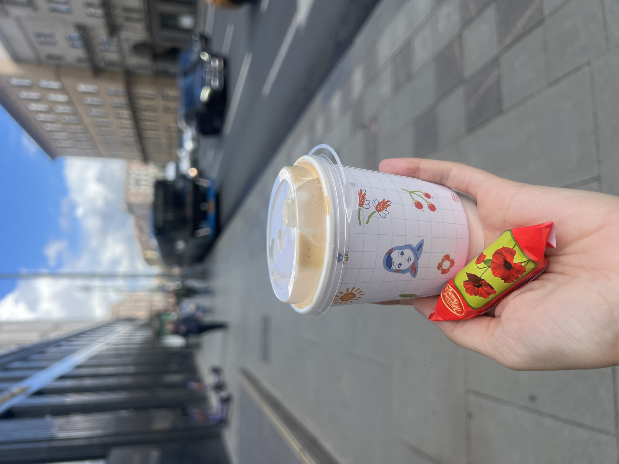 A hand holds a lidded cup and a wrapped candy on a city sidewalk. The cup is white with a grid pattern and decorated with small images of flowers, a sun, and a girl's face. The candy wrapper is red with a green background and an image of red poppies. In the background, there's a street with vehicles and buildings. The photo was likely taken in Moscow, Russia, as the text on the candy wrapper reads "Красный Октябрь Москва."