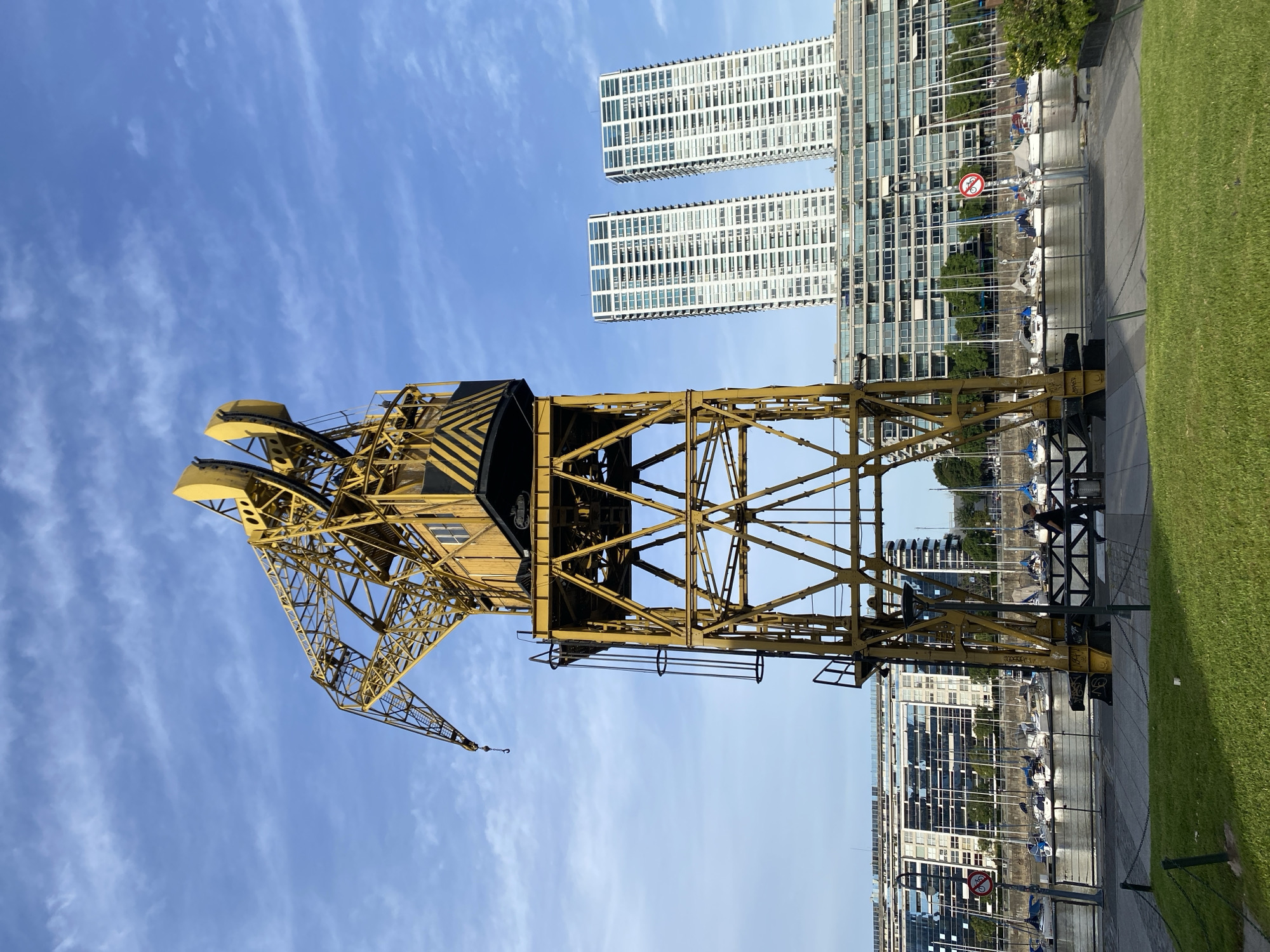 The image depicts a large yellow industrial crane in the foreground, with the city of Buenos Aires, Argentina visible in the background. The crane is constructed of steel beams and features a black and yellow striped section. Two modern high-rise buildings with numerous windows are prominent behind the crane. A waterfront area with boats is visible between the crane and the buildings. The sky is blue with scattered clouds. Green grass is visible on the right edge of the image. A small speed limit sign is seen near the edge of the water.