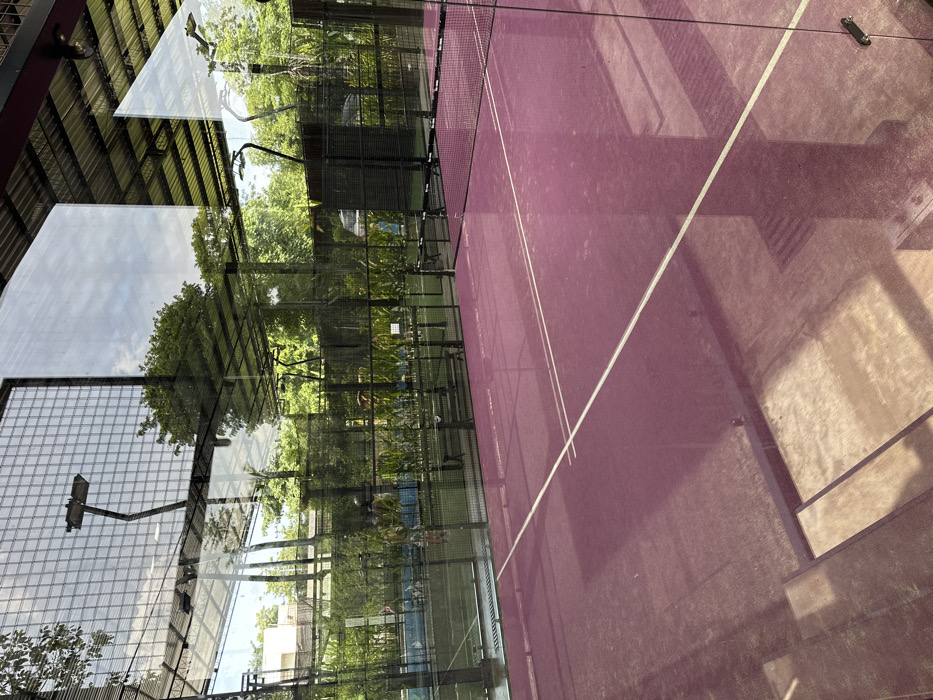 A sideways view of a purple padel court with white lines. Trees and vegetation are visible through the glass wall surrounding the court. Reflections of the sky and other buildings are superimposed on the glass. The court's net divides the space. Parts of the court are in shadow, adding texture to the image.