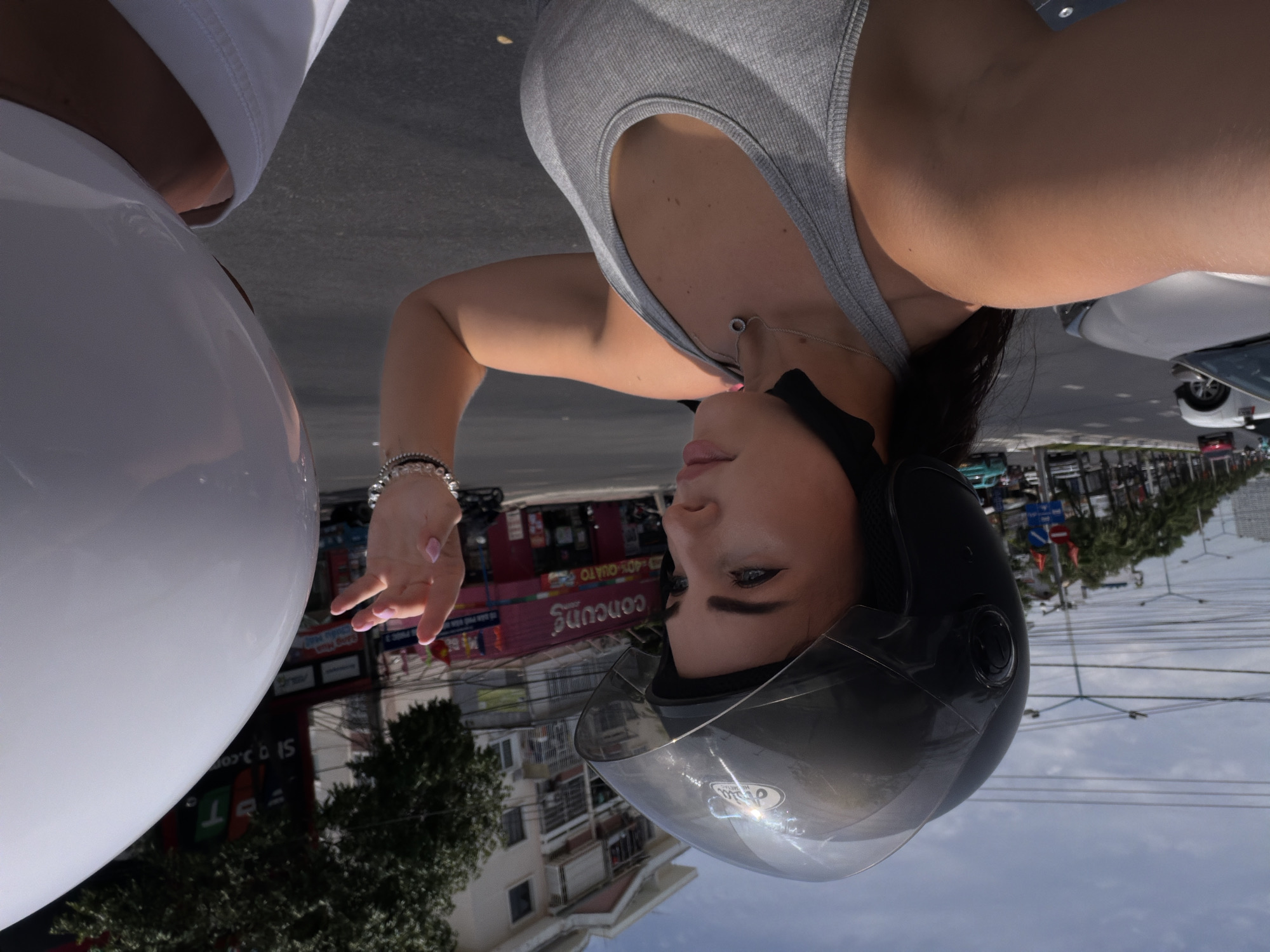 A medium shot shows a person wearing a gray tank top and a helmet. The person has black hair, light skin, and manicured nails. They are sitting on a white scooter. A street in Bac Nha Trang Ward, Vietnam, is in the background, with buildings, trees, cars, and power lines. The sky is partly cloudy.