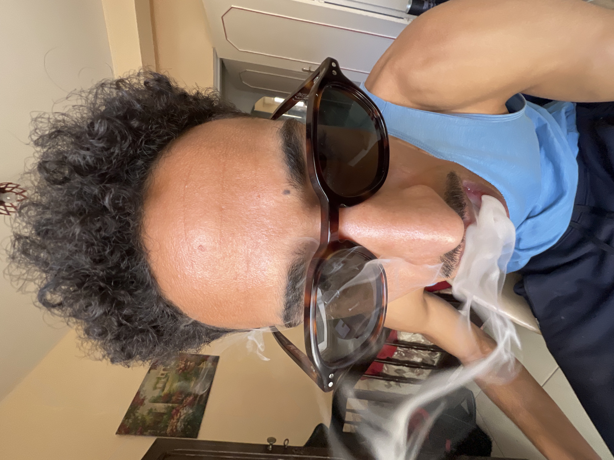 A close-up, wide-angle, frontal photograph depicts an individual with dark, curly hair and tanned skin, exhaling a significant plume of white vapor from their mouth. The individual wears brown tortoise-shell sunglasses, partially obscuring the eyes and the upper portion of the nose. A faint mustache is visible above the vapor. A small dark mole is present on the forehead. The individual wears a light blue tank top. The background features light beige walls, a framed landscape painting on the left wall, and a decorative red-toned light fixture partially visible on the ceiling. To the right, a white two-panel door with dark trim is slightly ajar, revealing an interior space. A light-colored tiled floor is visible in the lower left. The scene is an indoor environment located in the city of Tanger, Morocco.